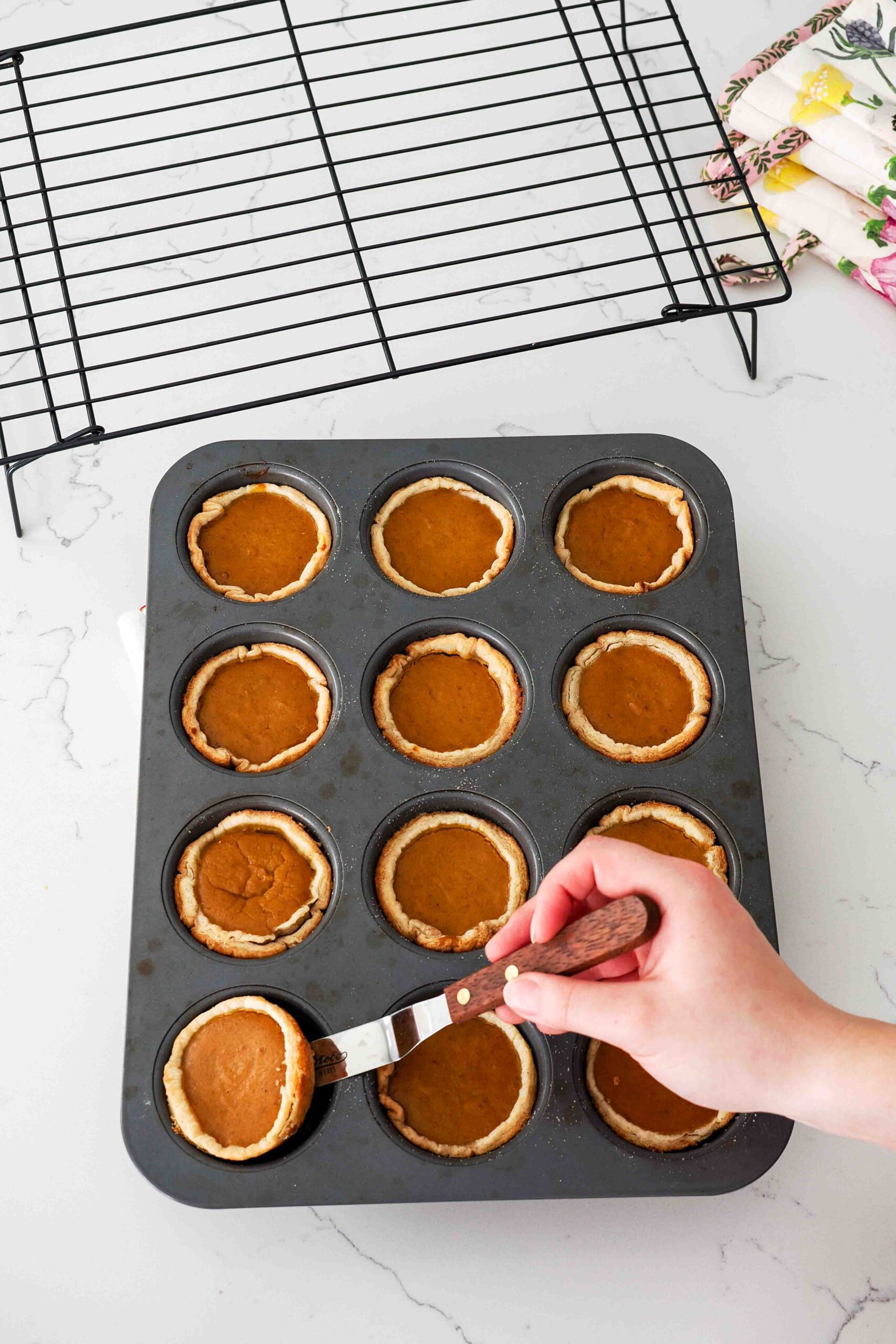A hand uses an offset spatula to remove a mini pumpkin pie from a muffin pan hole.