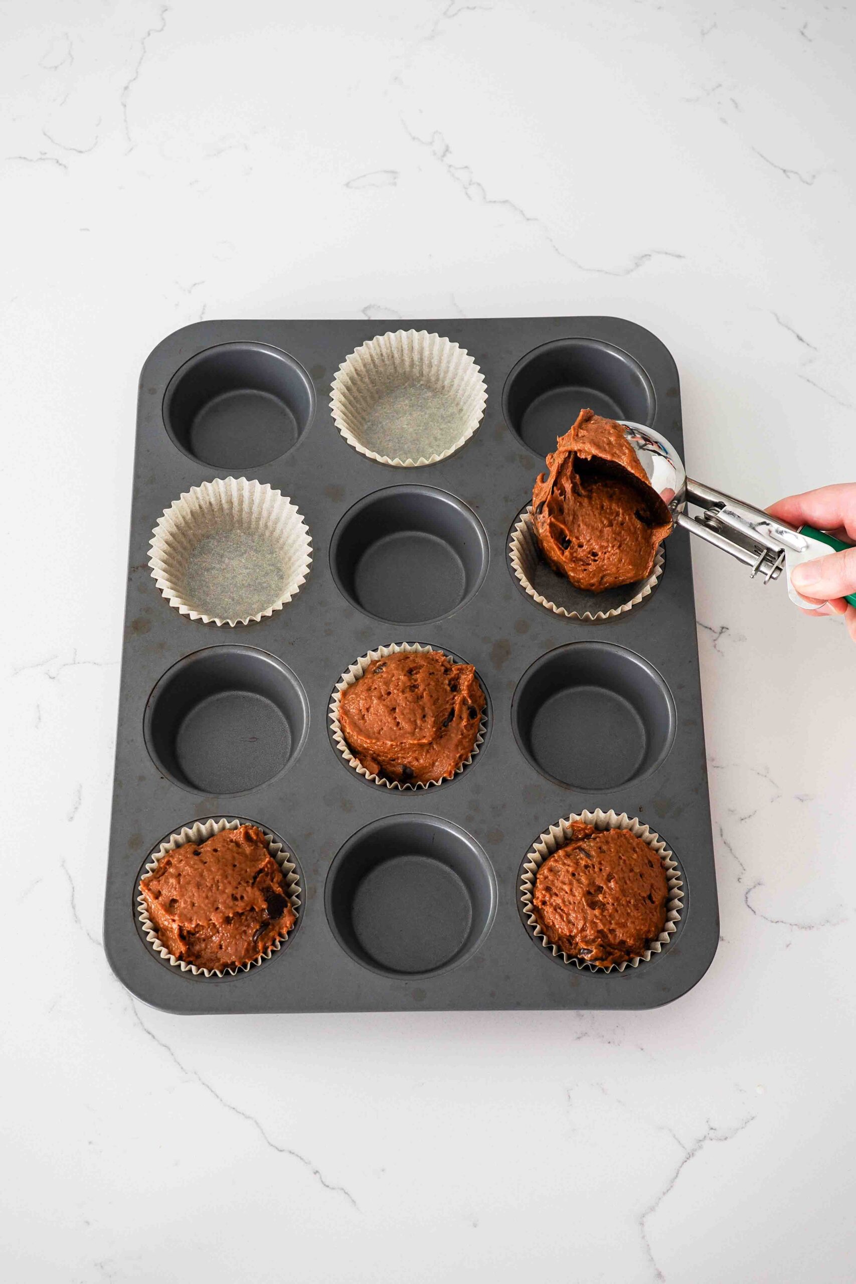 A hand scoops chocolate pumpkin muffin batter into a lined muffin pan.