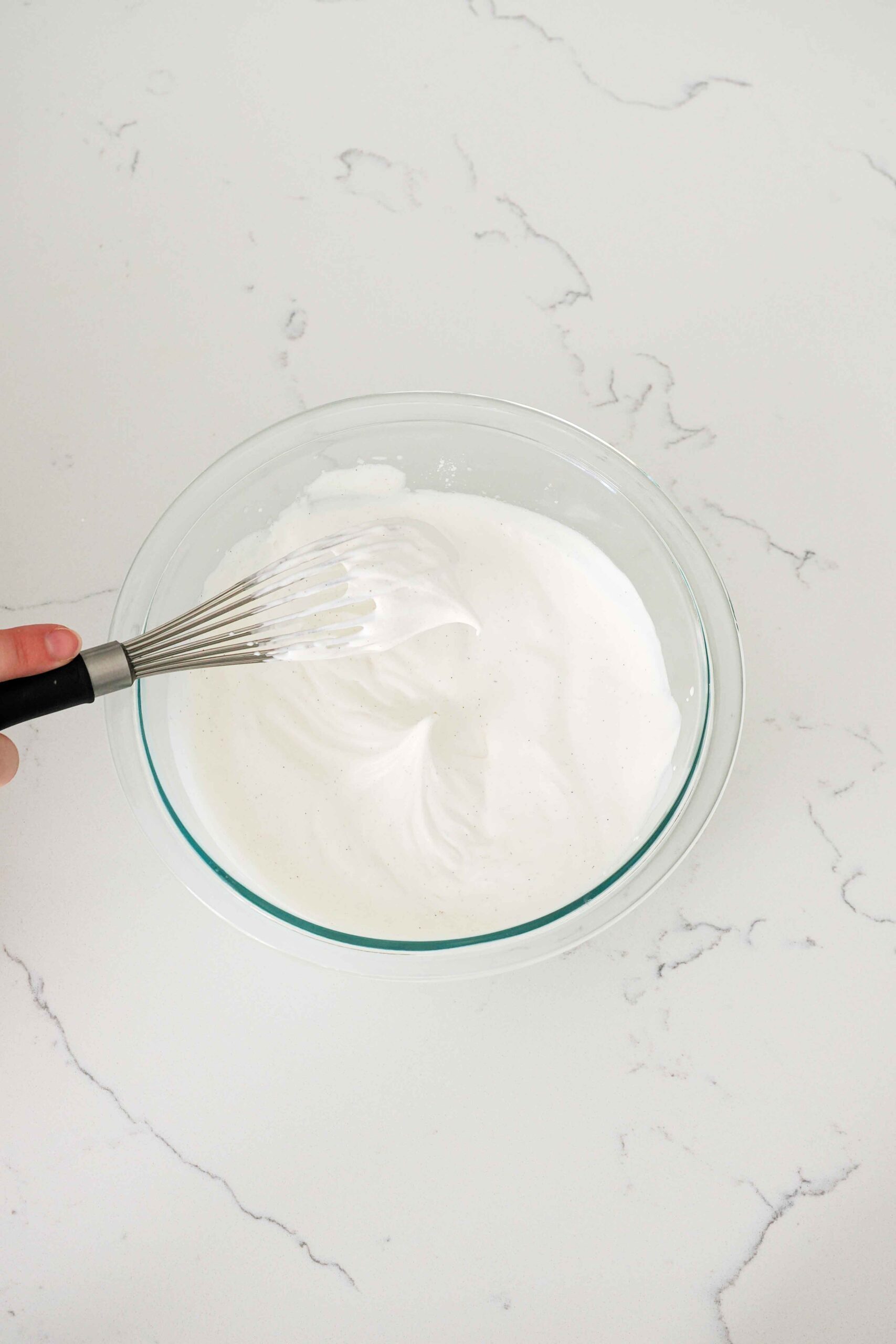 A whisk with a clump of whipped cream drooping off of it in soft peaks.