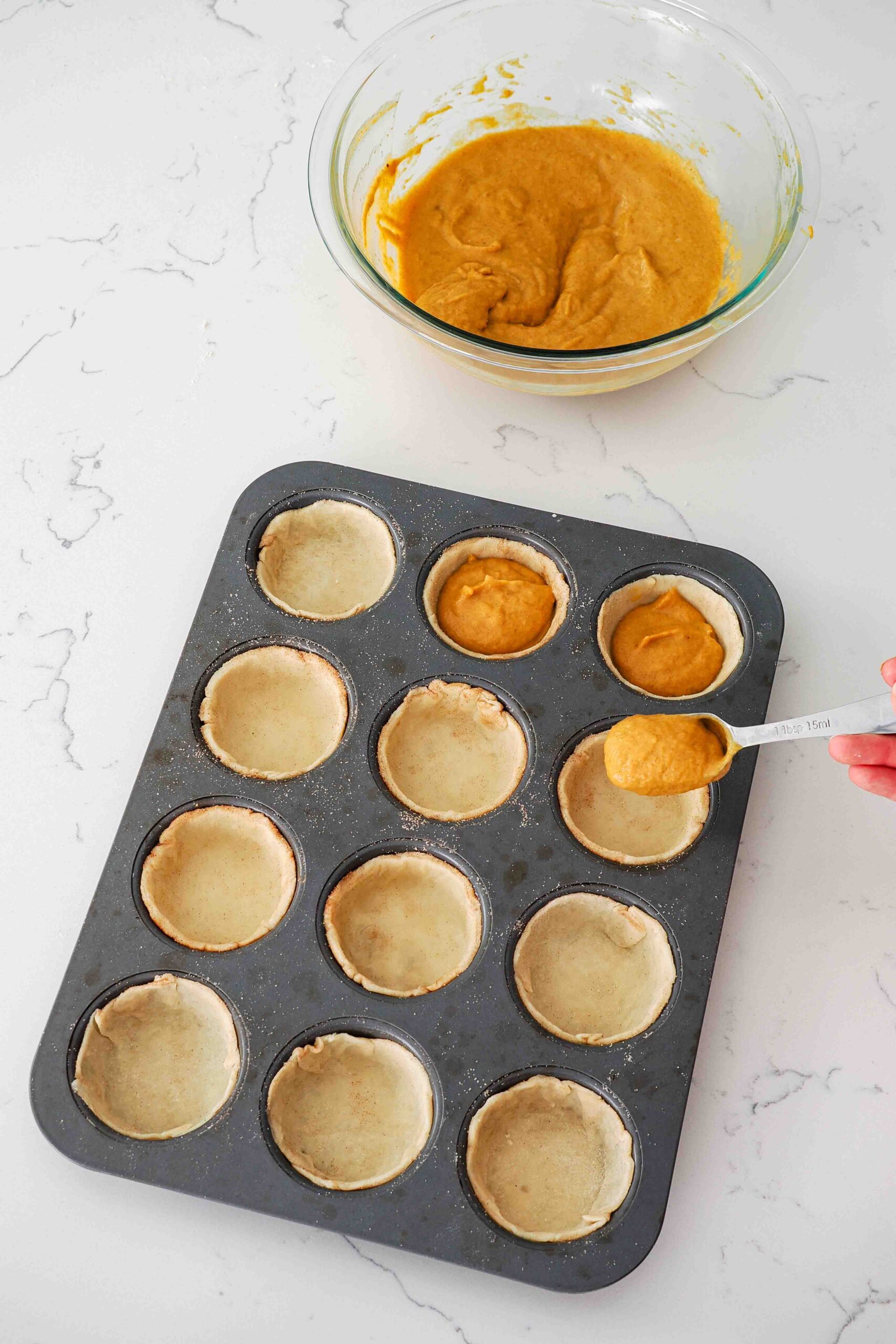 A tablespoon adds pumpkin pie filling to pie crusts in the holes of a muffin pan.