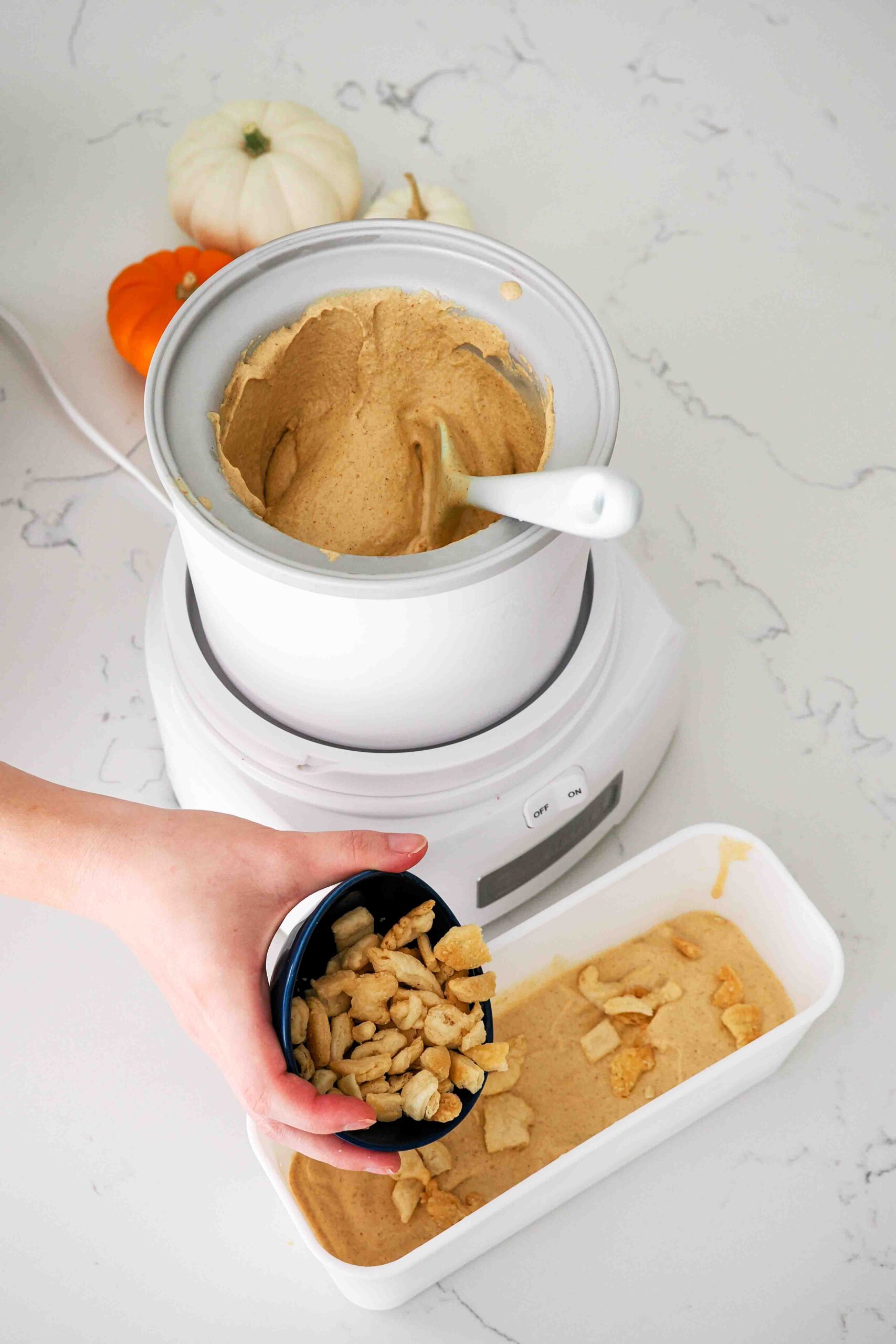 A hand sprinkles pie crust pieces into a container of homemade pumpkin ice cream.