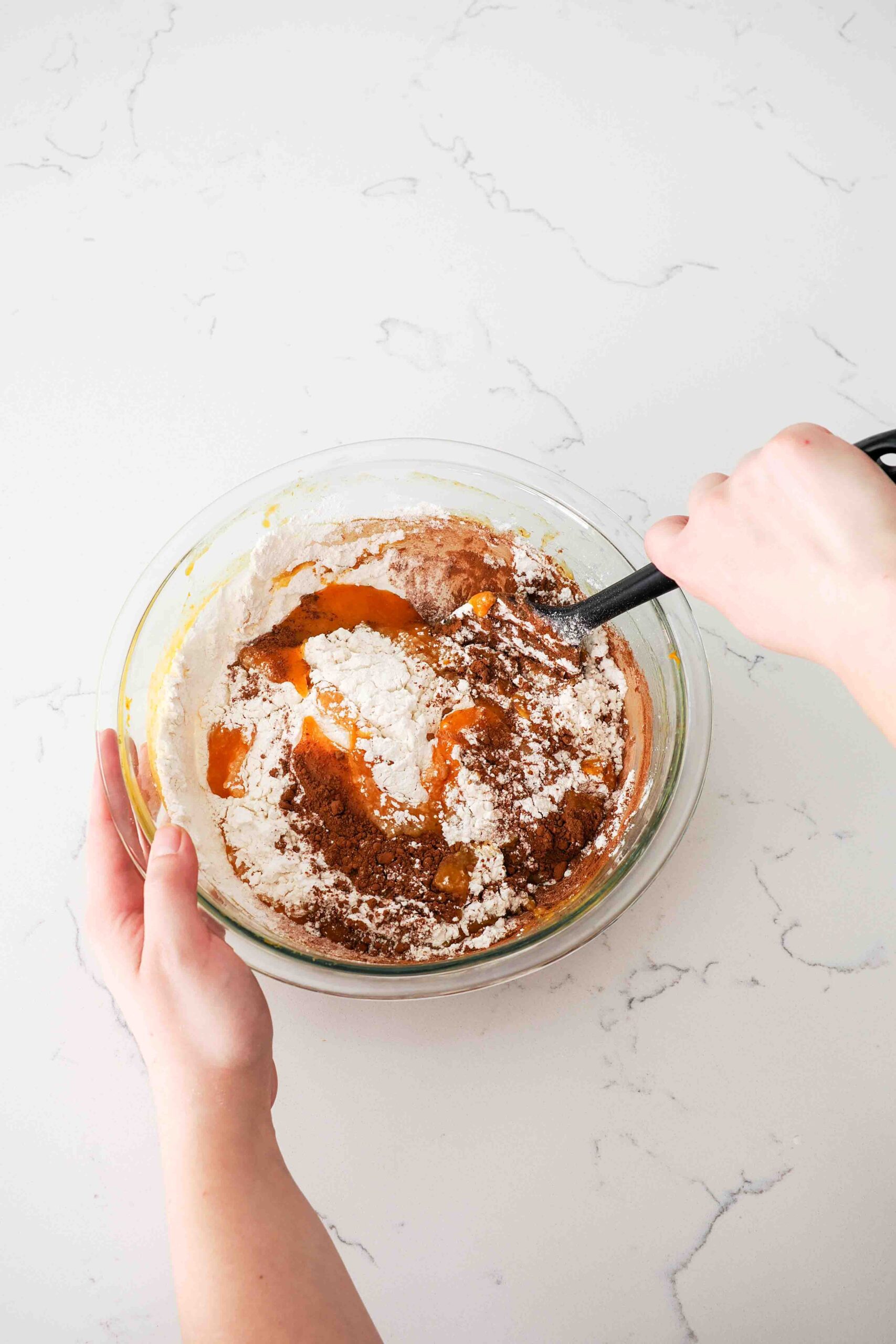 Two hands use a spatula to stir in the dry ingredients into a wet pumpkin batter.