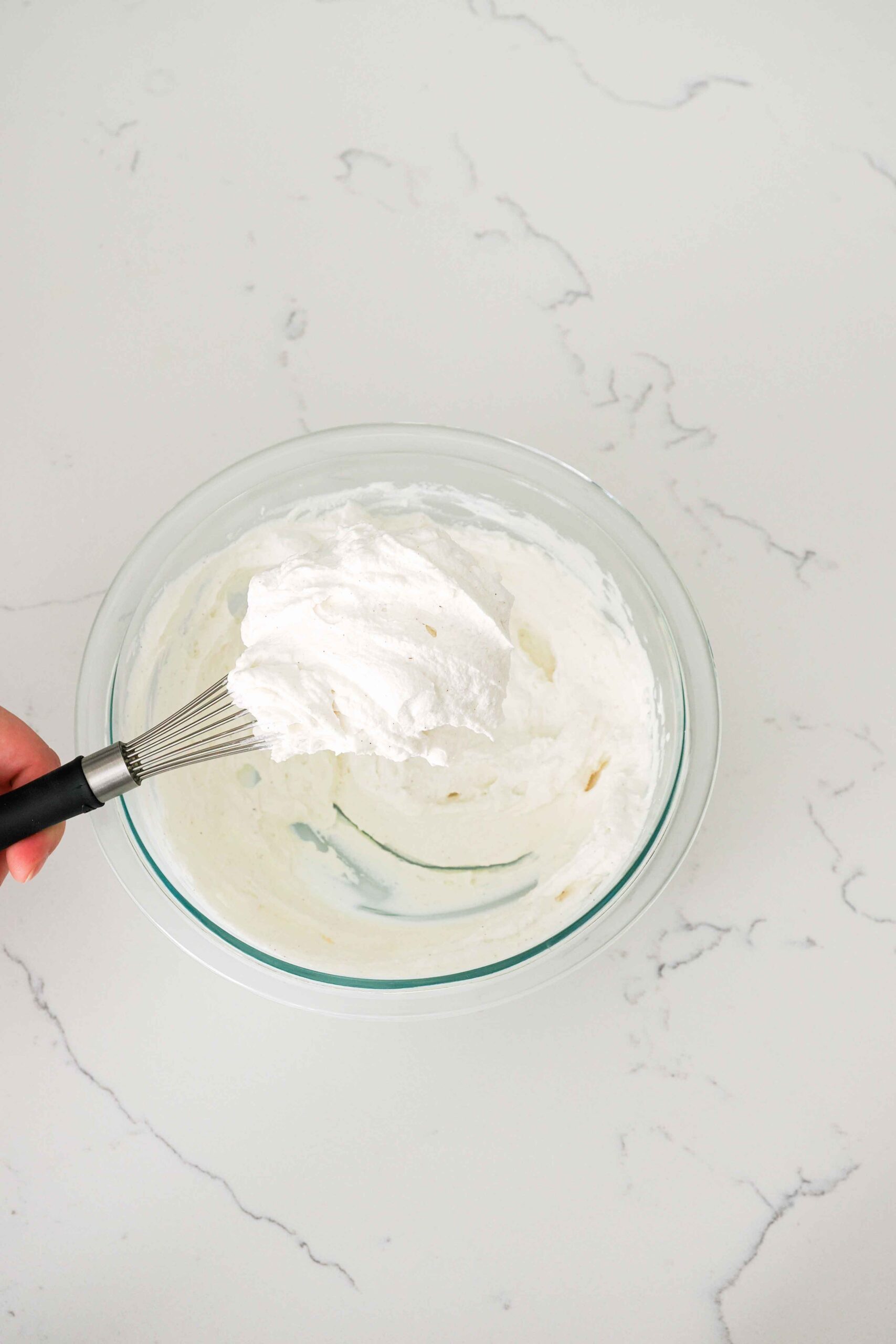 A whisk holds up a clump of whipped cream with stiff peaks.