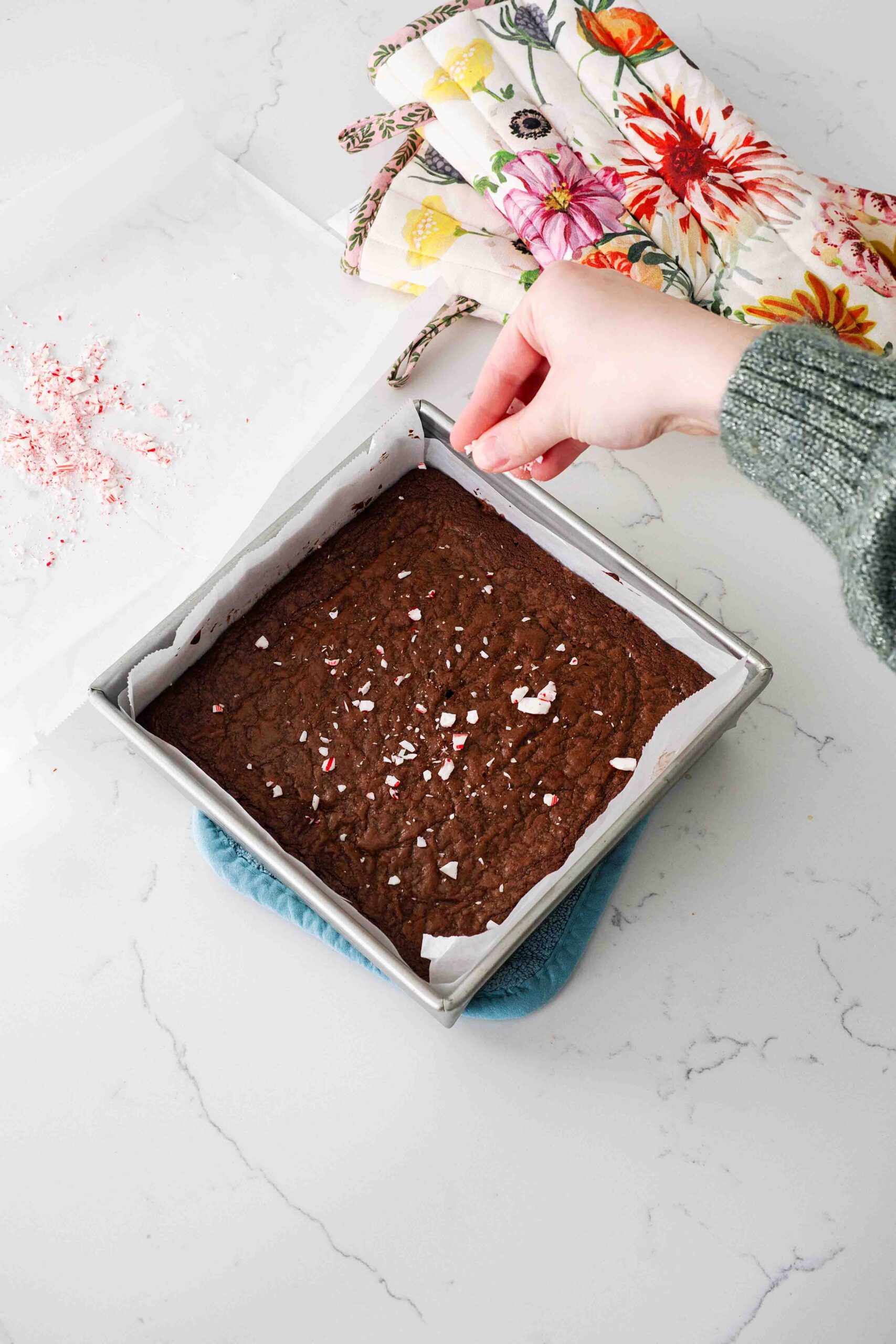 A hand sprinkles crushed candy canes over a hot pan of brownies.