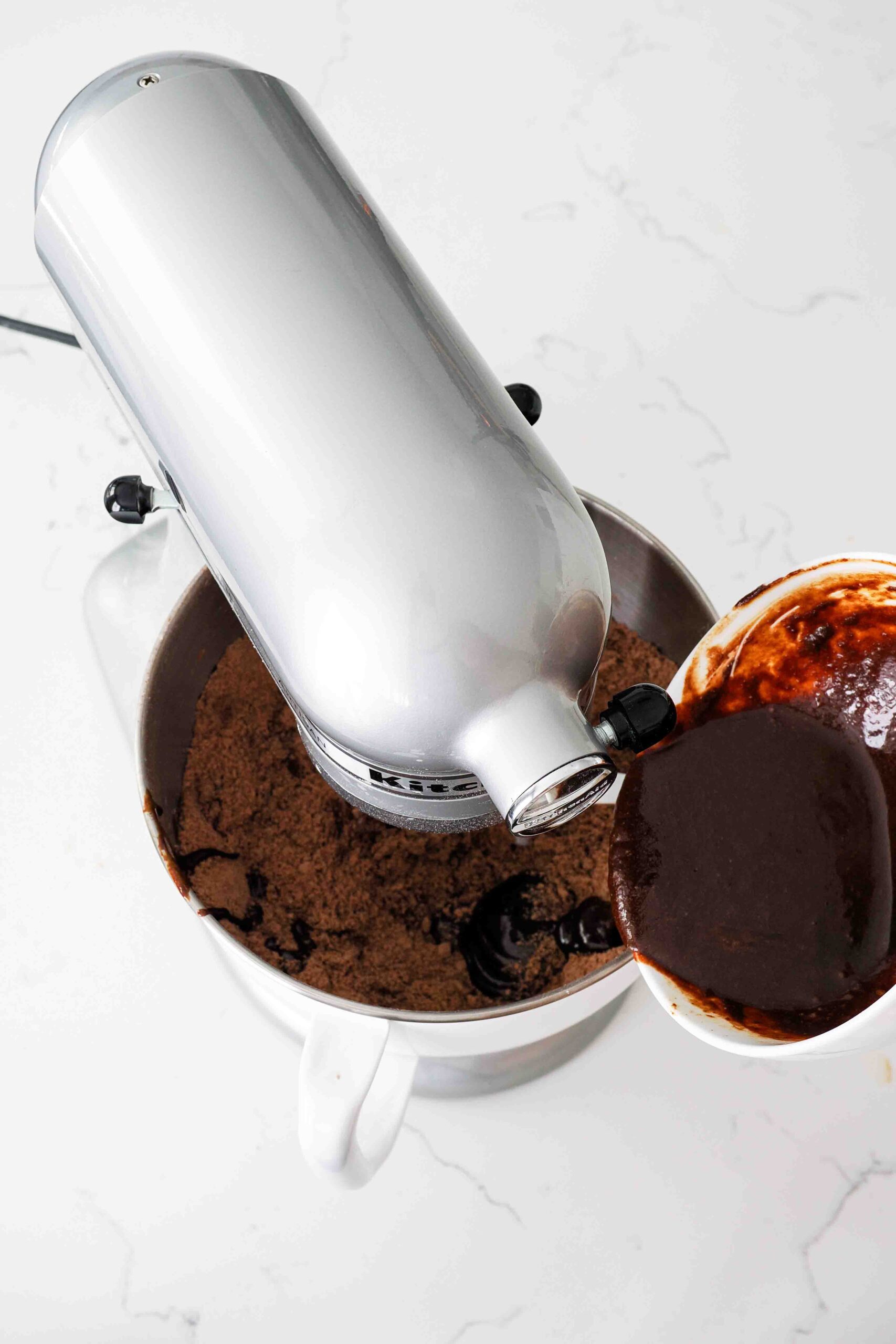 Dark liquid is poured into the bowl of a stand mixer with cocoa-colored dough.