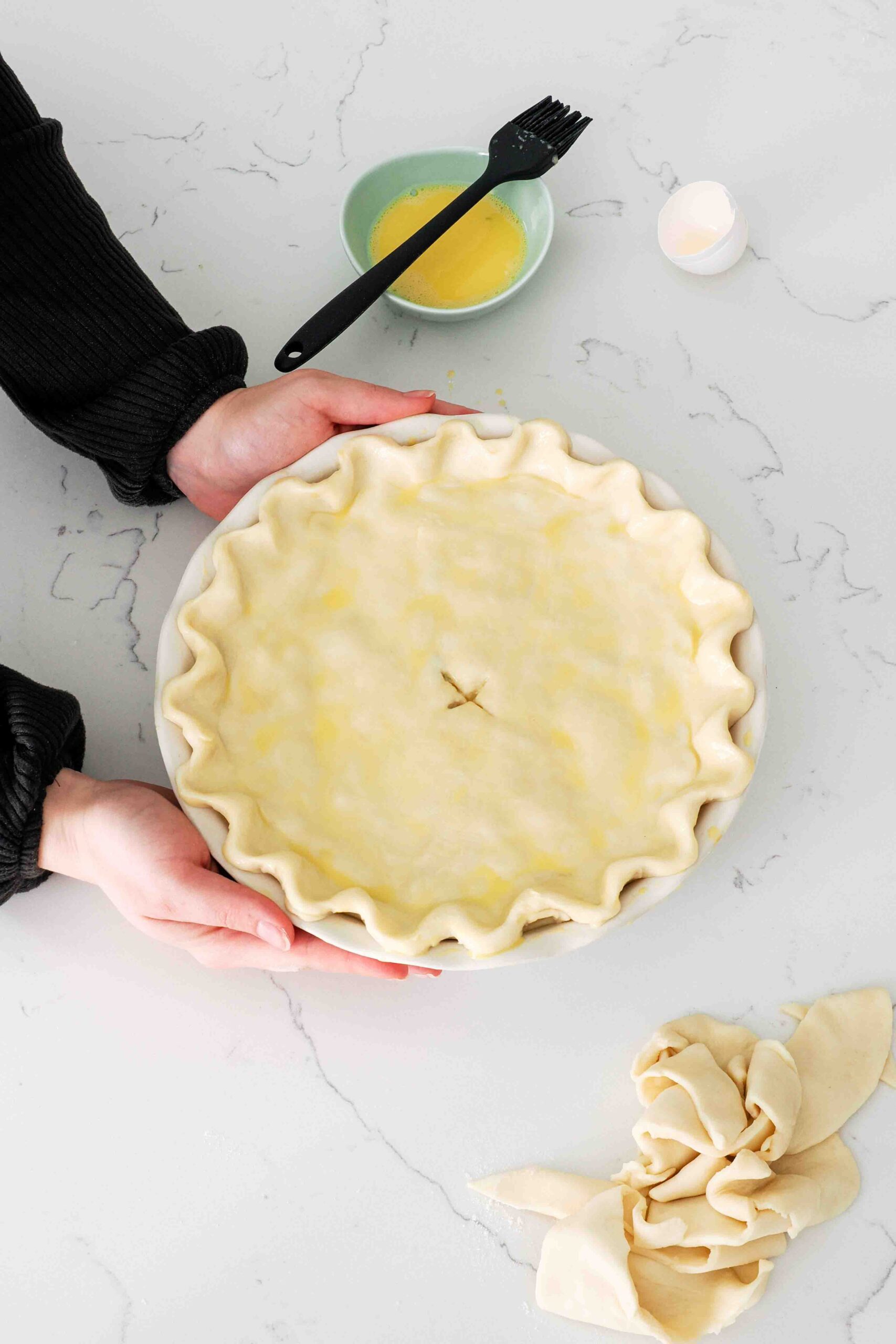 Two hands move an unbaked and egg-washed pie on a counter.