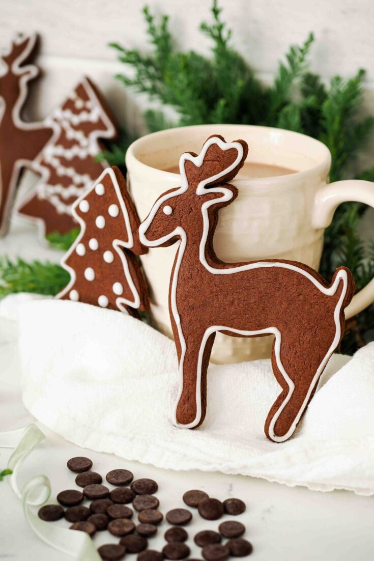 A chocolate gingerbread cookie in the shape of a reindeer resting against a cream-colored mug of hot cocoa.