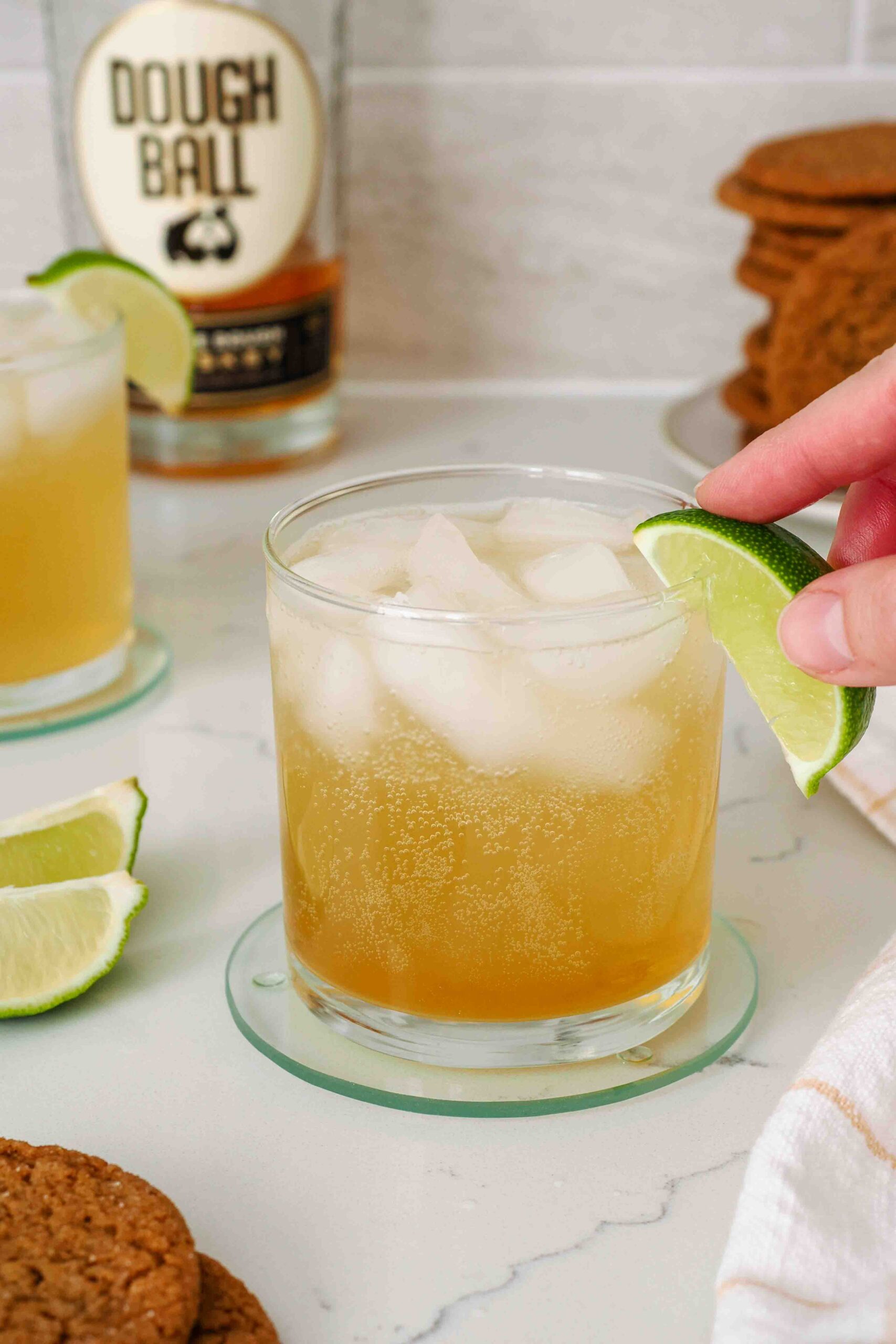 A hand adds a lime wedge to the side of a lowball glass with a whiskey gingersnap.