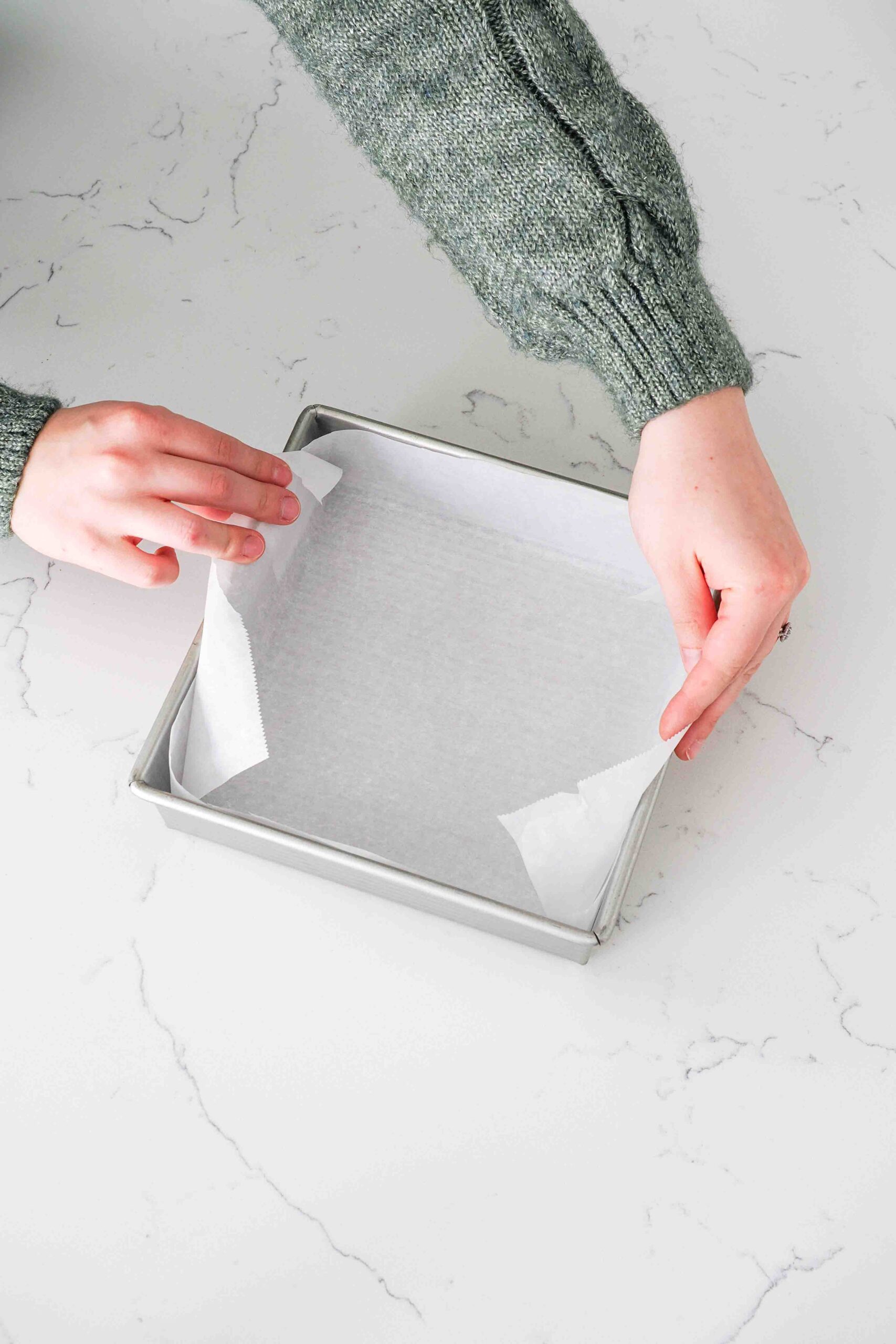 Two hands line a square metal pan with parchment paper.