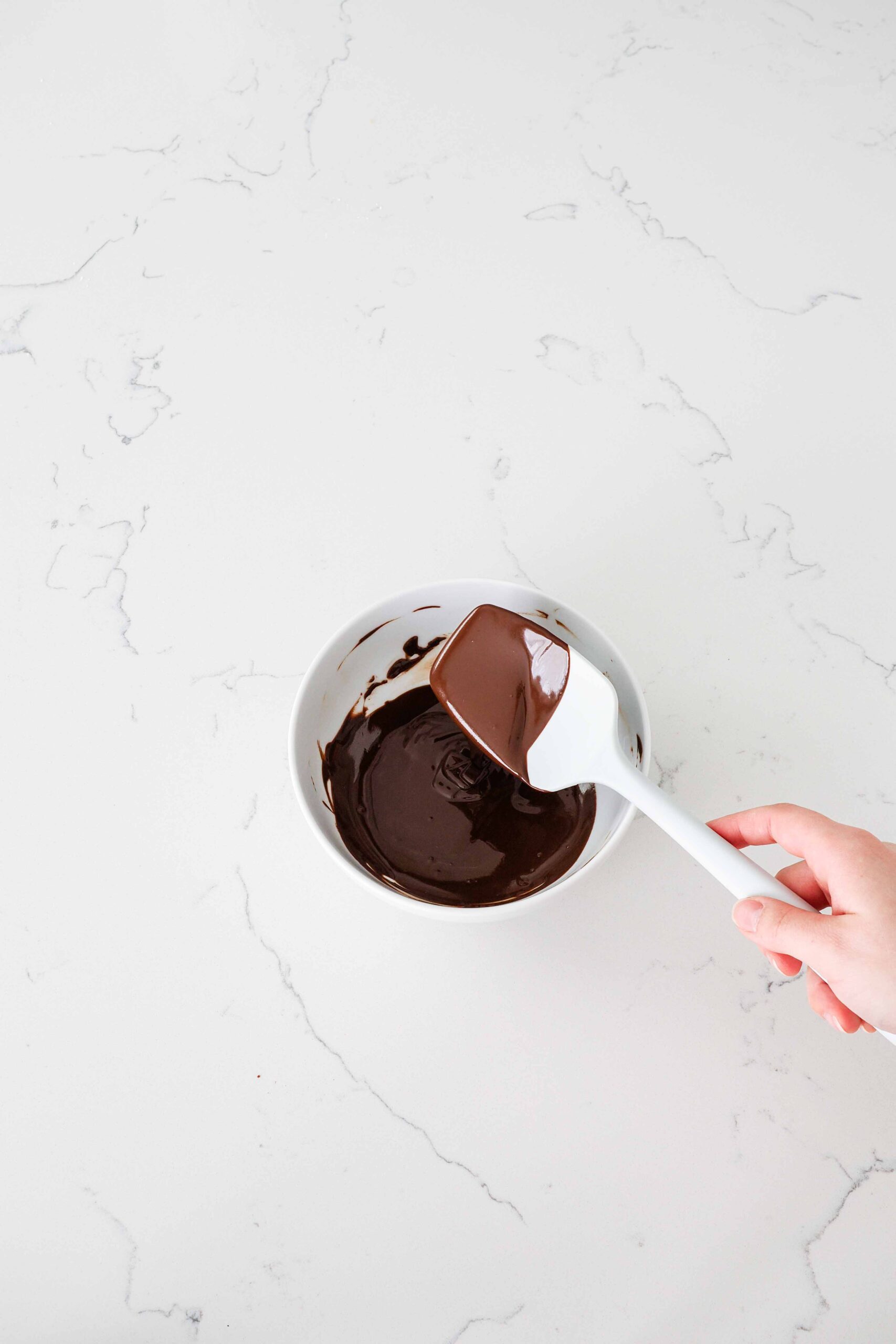A spatula with glossy, melted chocolate running off it into a bowl.