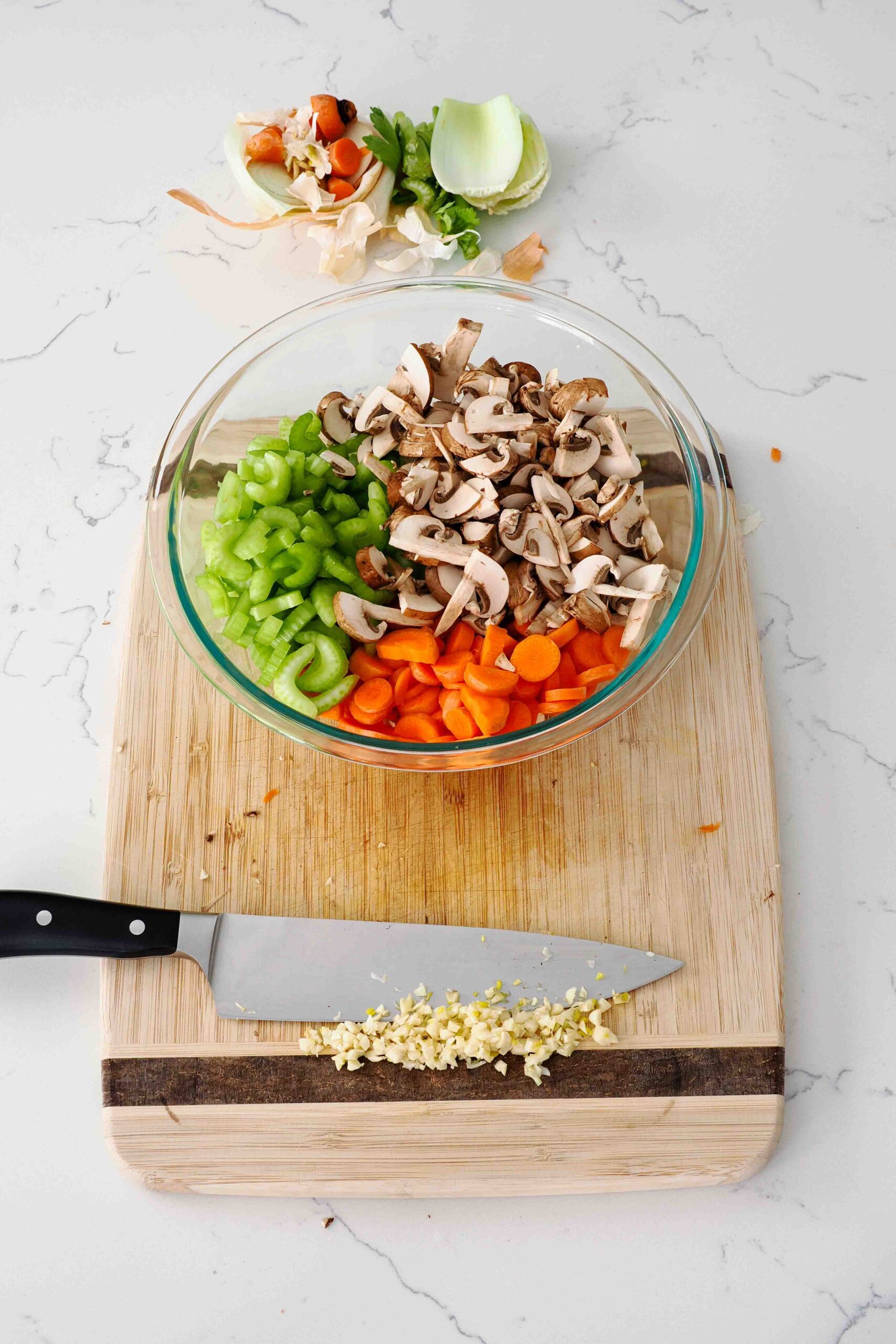 Chopped onion, celery, carrot, and mushrooms in a glass bowl, with minced garlic on a cutting board.