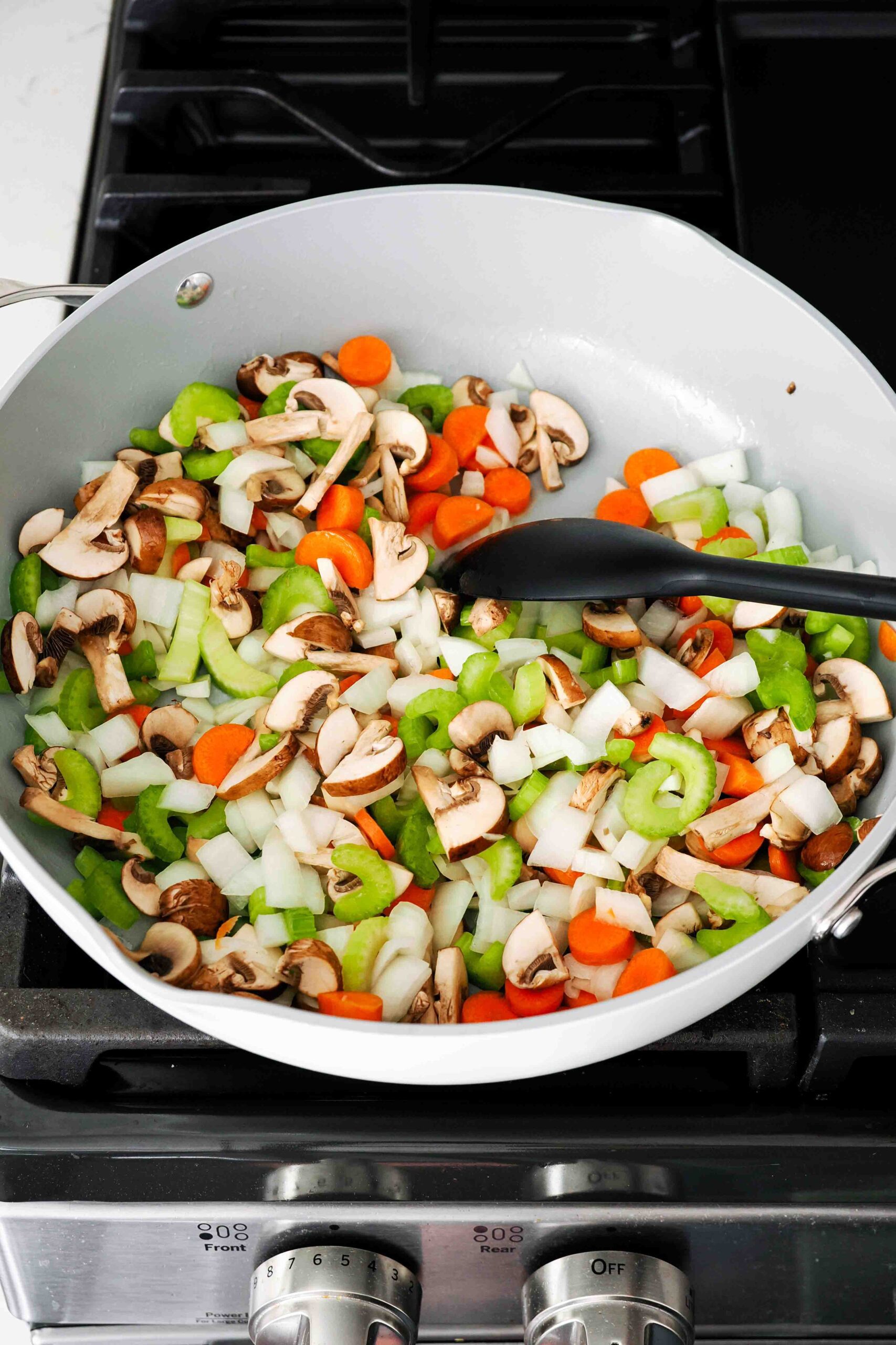 A spoon stirs diced onion, carrot, celery, and mushrooms in a pan with butter.