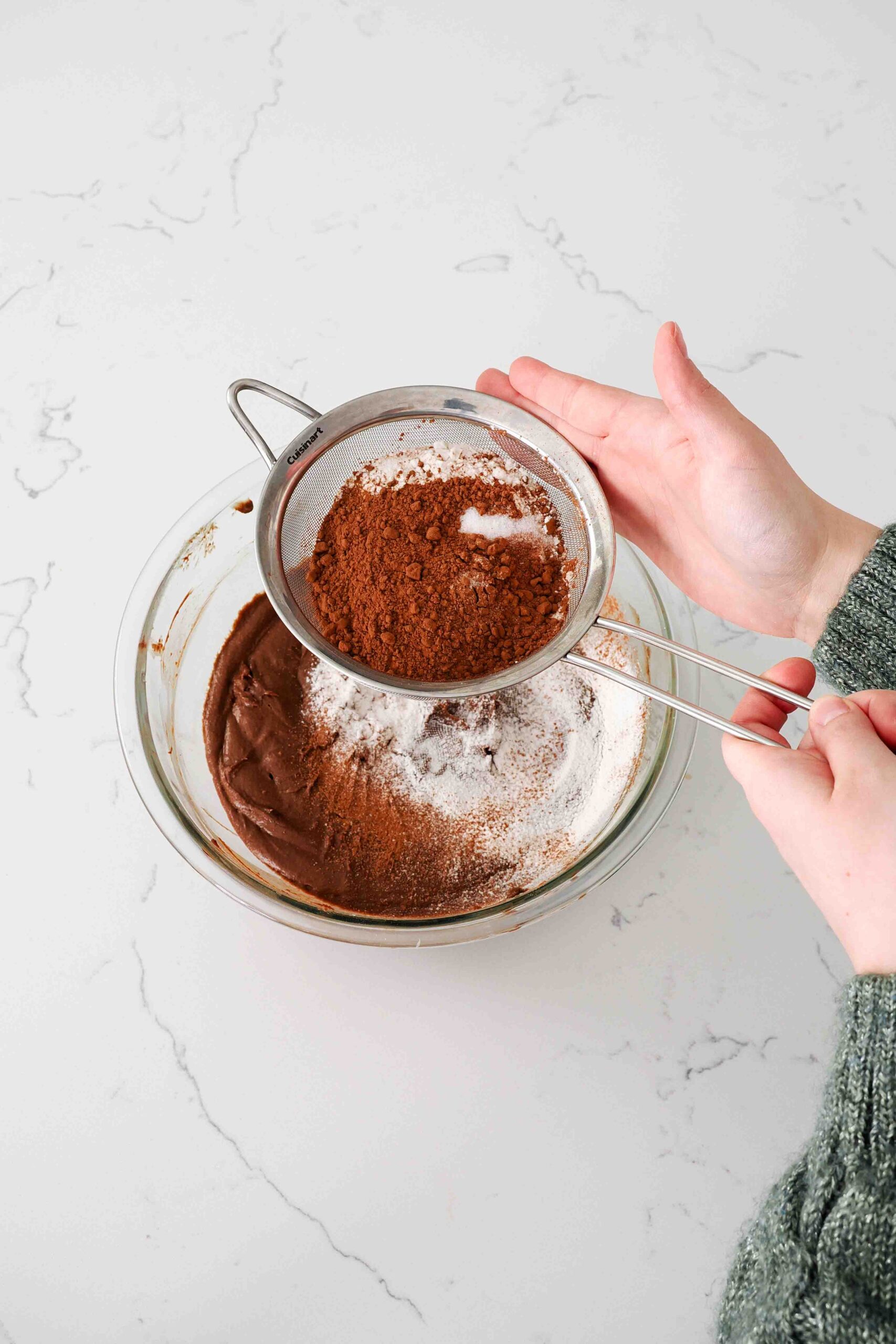 Two hands sift flour and cocoa powder into a glass bowl with brownie batter.
