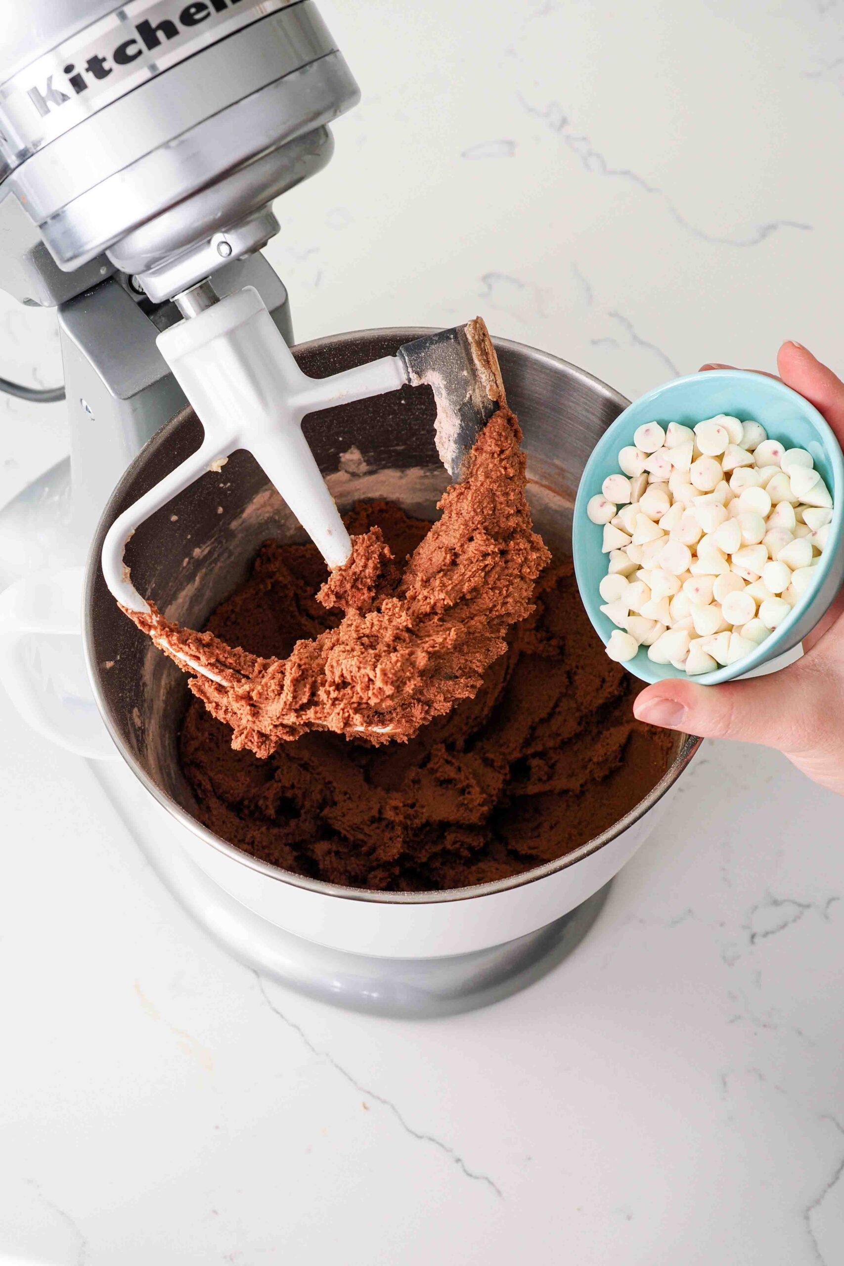 A hand adds peppermint chips into a mixing bowl with chocolate cookie dough.