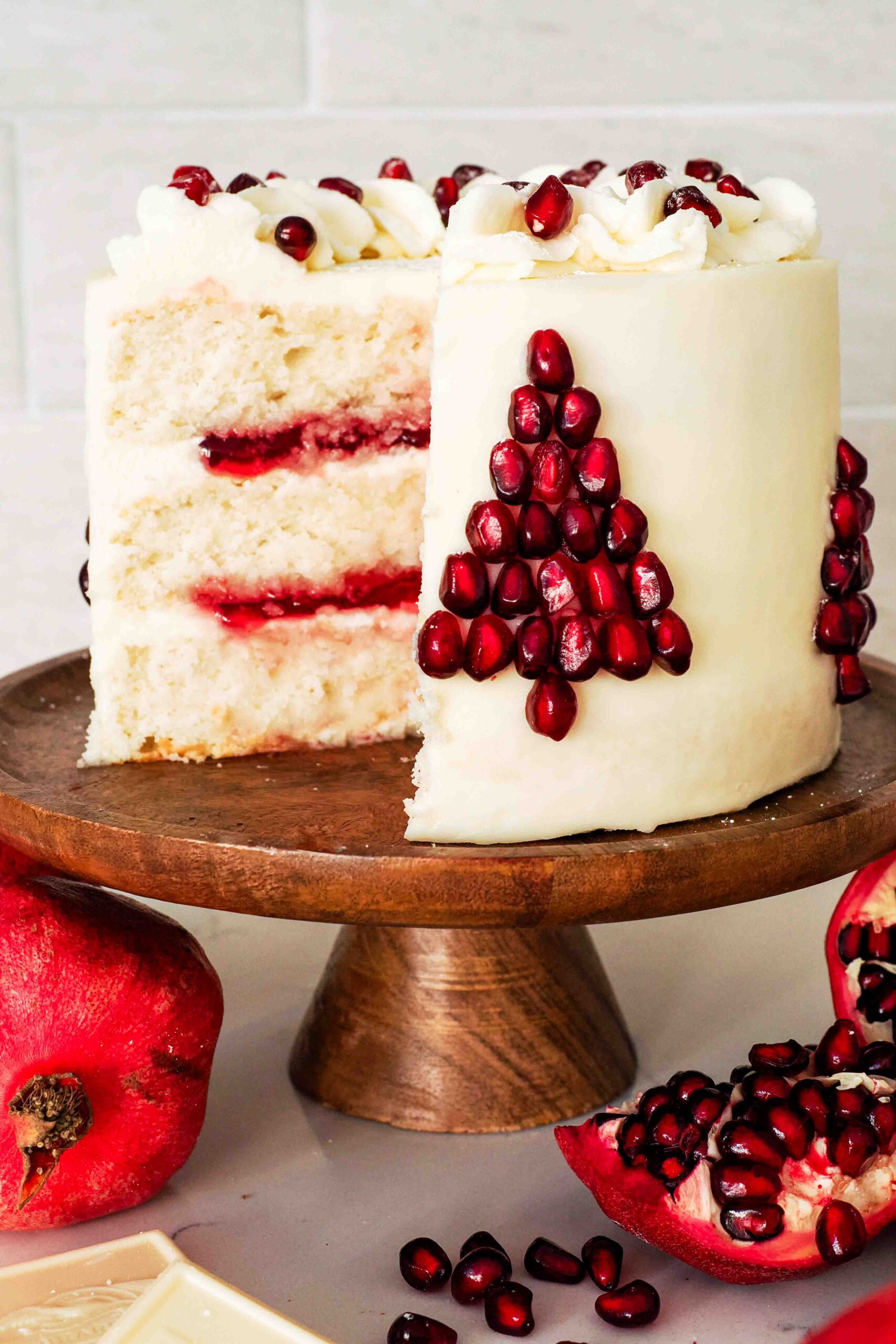 A white layer cake filled with pomegranate jelly, frosted with white chocolate buttercream, and decorated with pomegranate seeds in the shape of a Christmas tree.