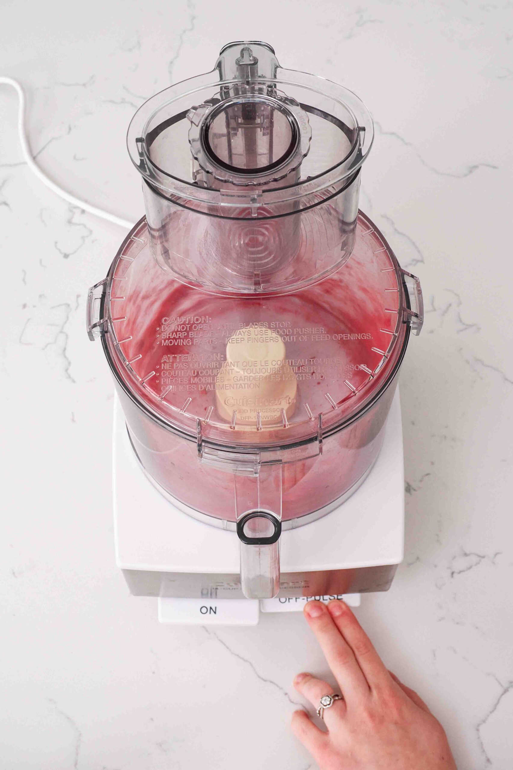 A hand presses the "pulse" button on a food processor with freeze-dried raspberries.