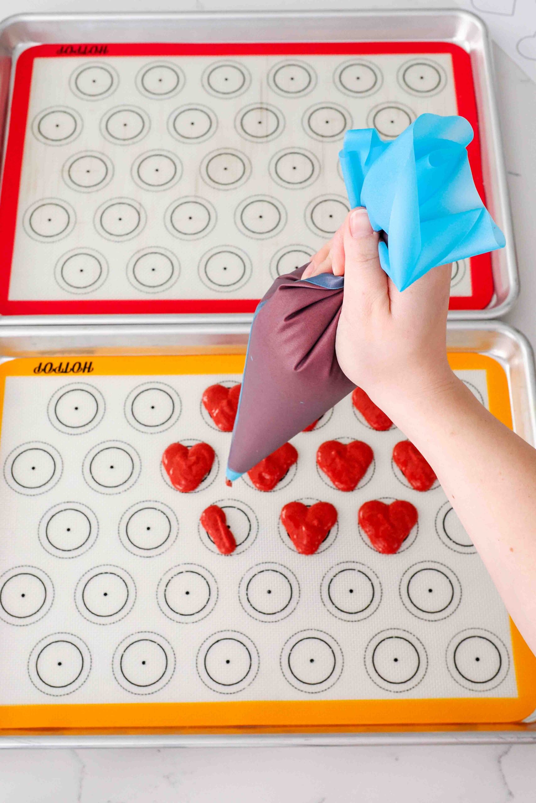 A hand pipes the left half of a heart-shaped macaron on silicone macaron mats.