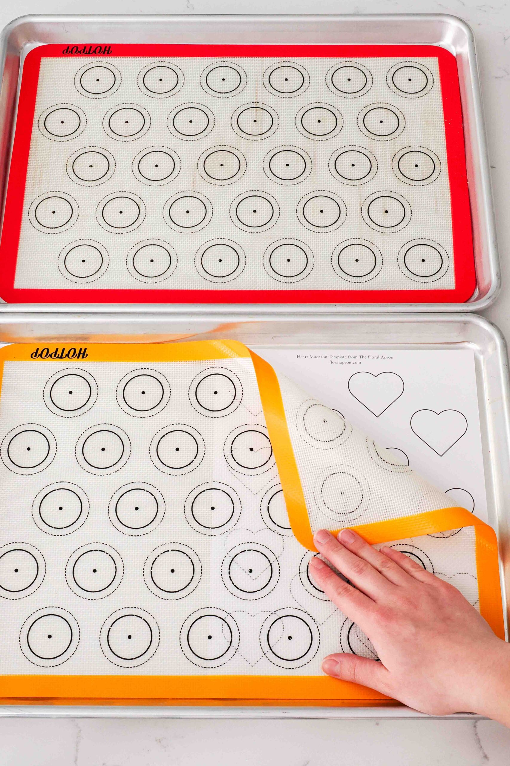 Two baking sheets with silicone macaron mats and a paper template with hearts underneath.