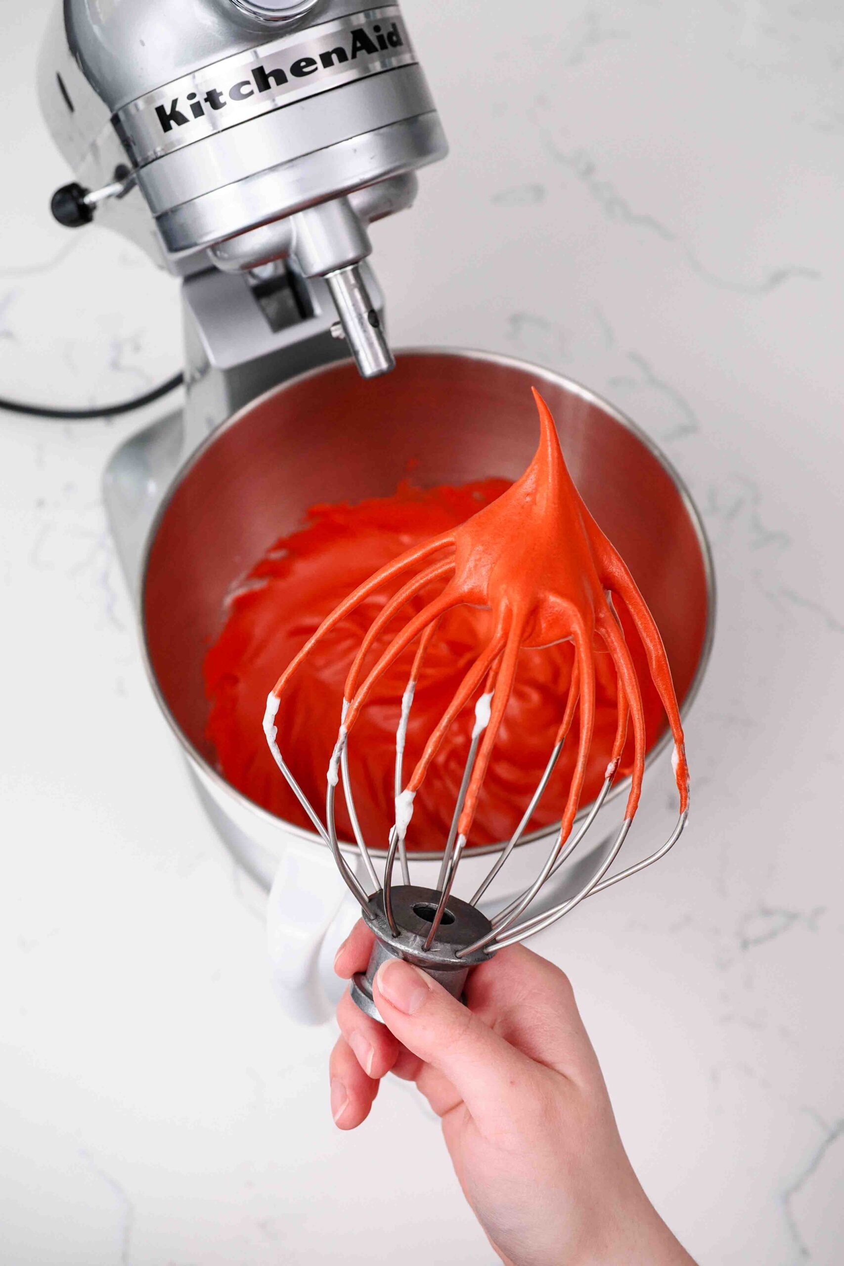 A hand holds up a whisk with a stiff peak of red meringue pointing up.