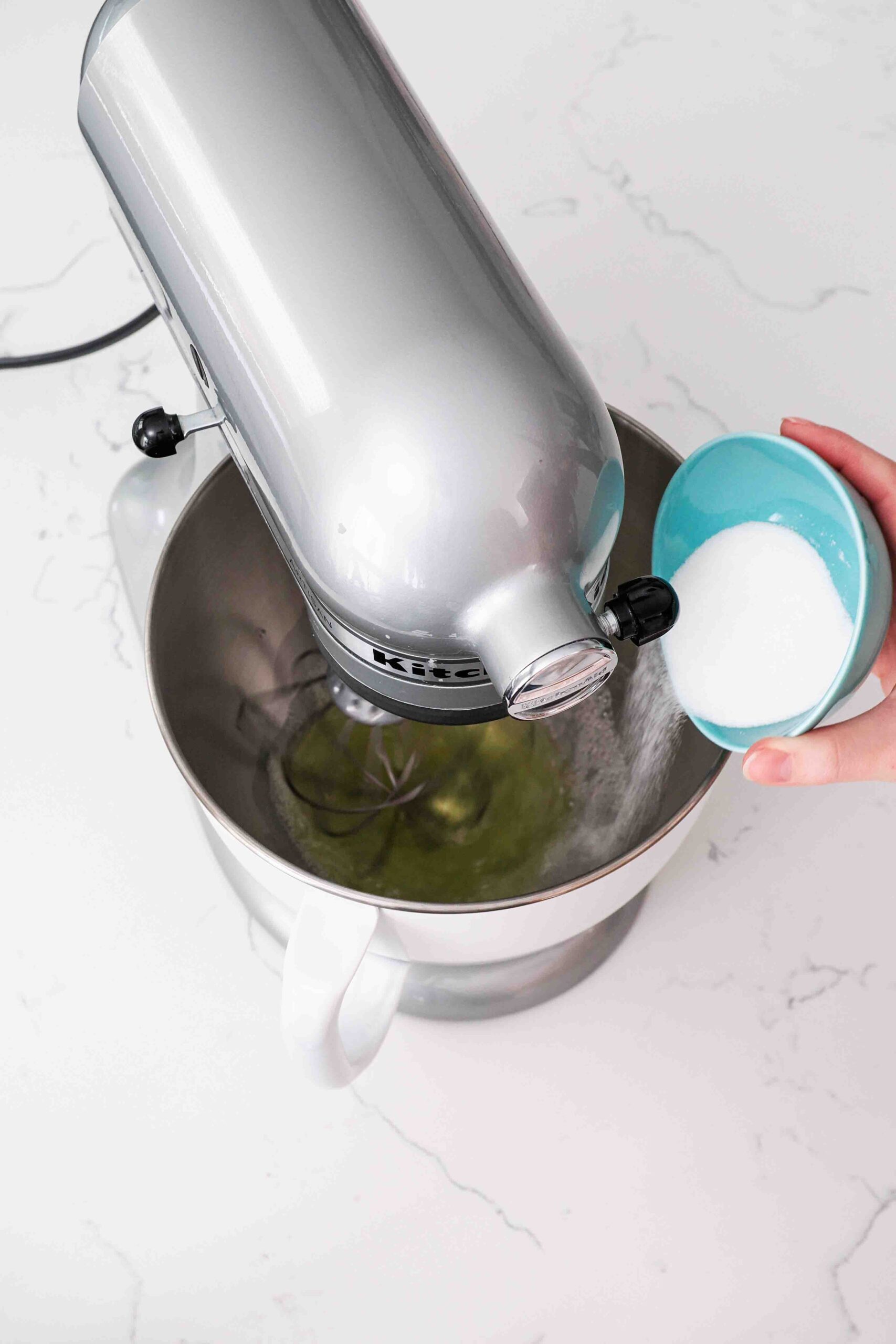A hand sprinkles granulated sugar into the bowl of a stand mixer whisking egg whites.