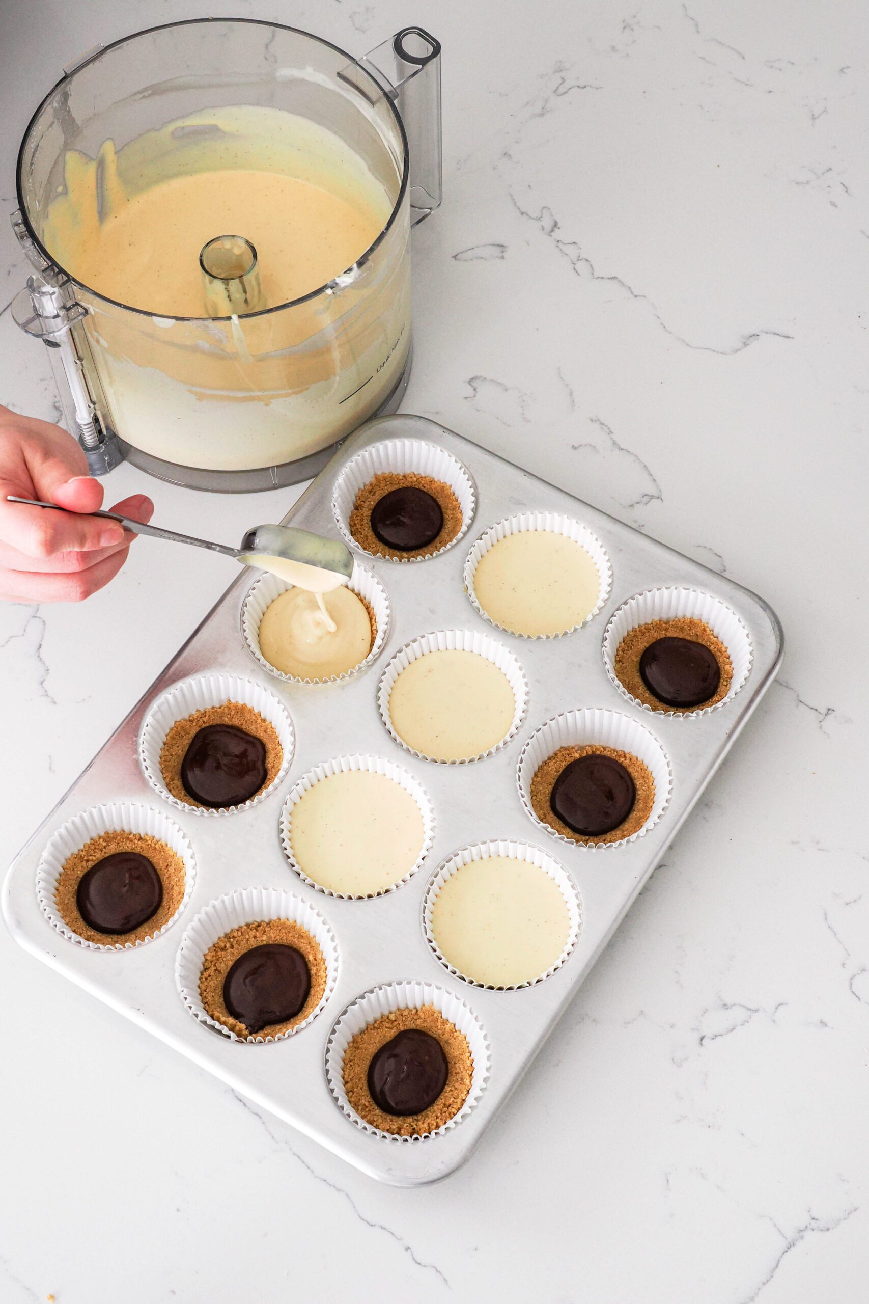 A hand drops a Tablespoon of cheesecake filling into muffin liners at a time.
