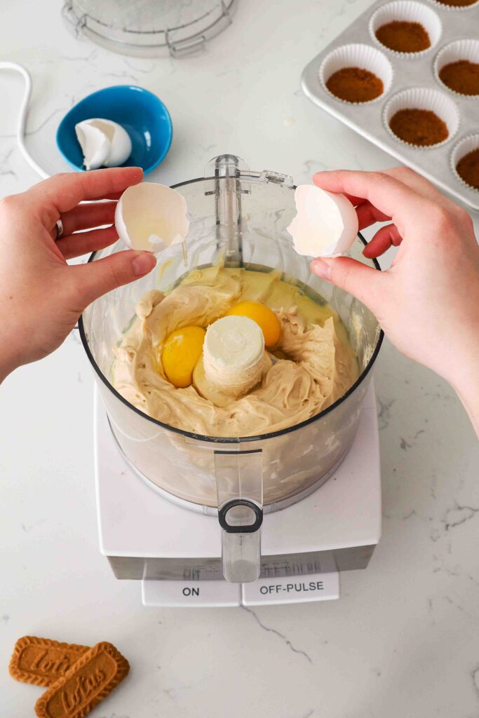 Two hands break an egg into the bowl of a food processor with Biscoff cheesecake filling.