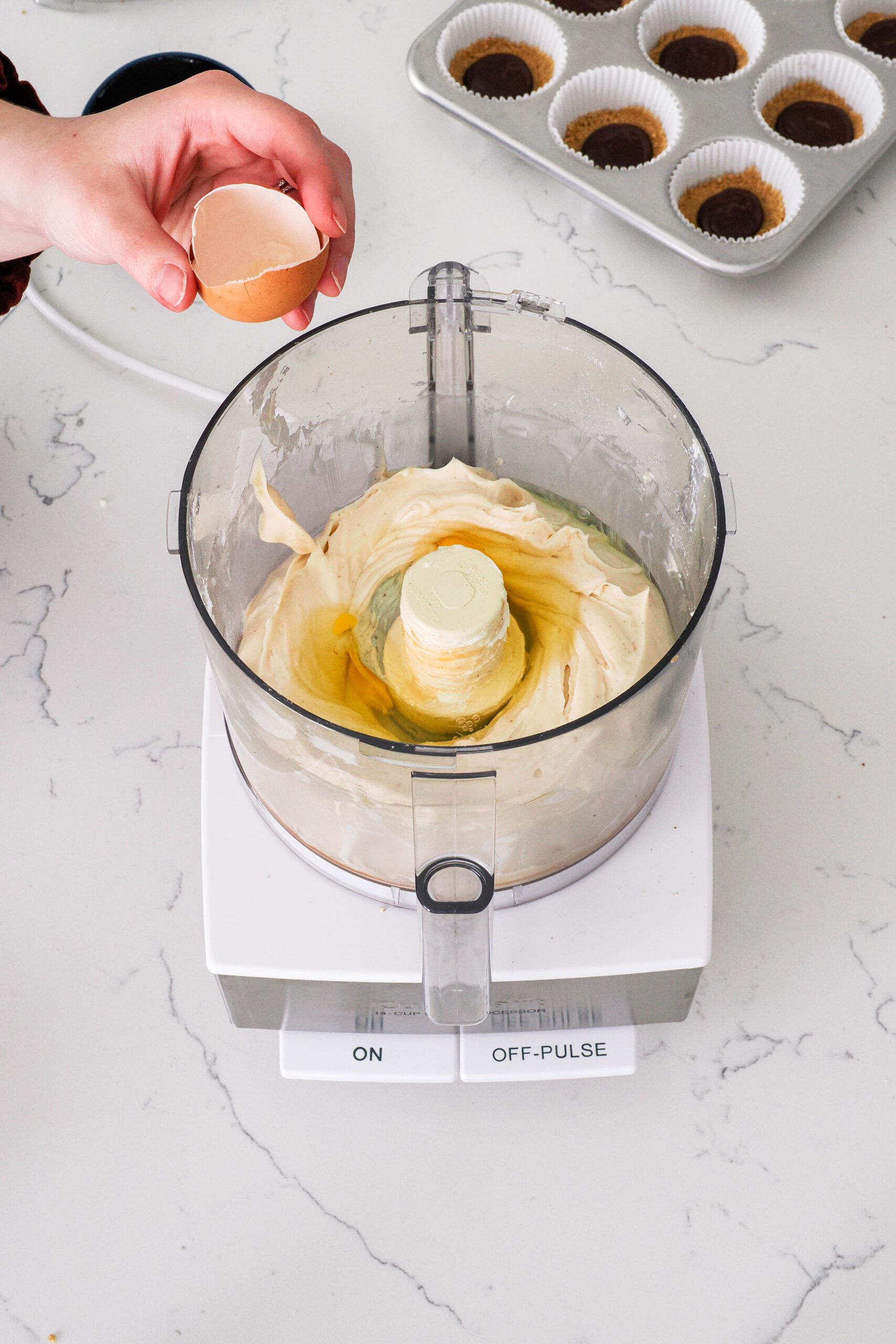 A hand cracks eggs into the bowl of a food processor with caramel cheesecake filling.
