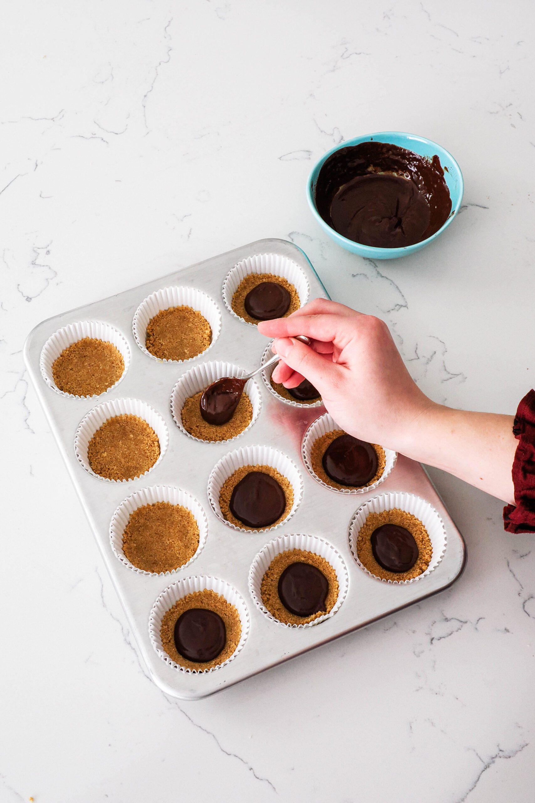 A hand adds a teaspoon of ganache on top of graham cracker crusts in paper muffin liners.