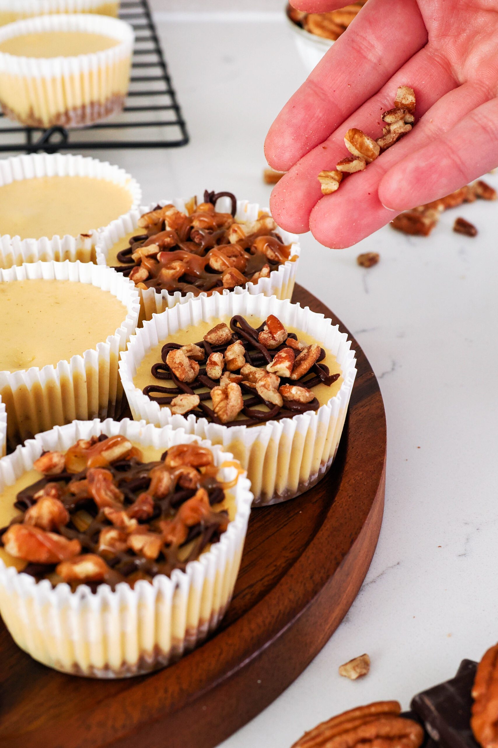 A hand adds pecan pieces to the top of a mini turtle cheesecake.
