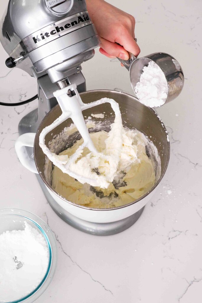 A hand adds powdered sugar a cup at a time into a stand mixer bowl with butter.