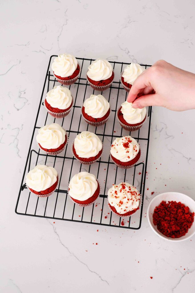 A hand sprinkles red velvet crumbs on top of frosted red velvet cupcakes.