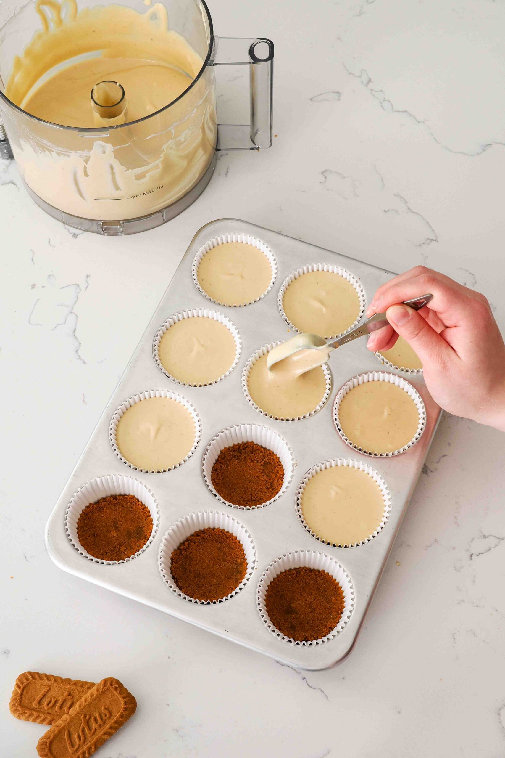 A hand drops a Tablespoon of Biscoff cheesecake filling into paper liners in a muffin pan at a time.