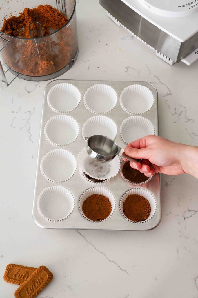 A hand uses a measuring cup to flatten cookie crusts in a muffin pan.
