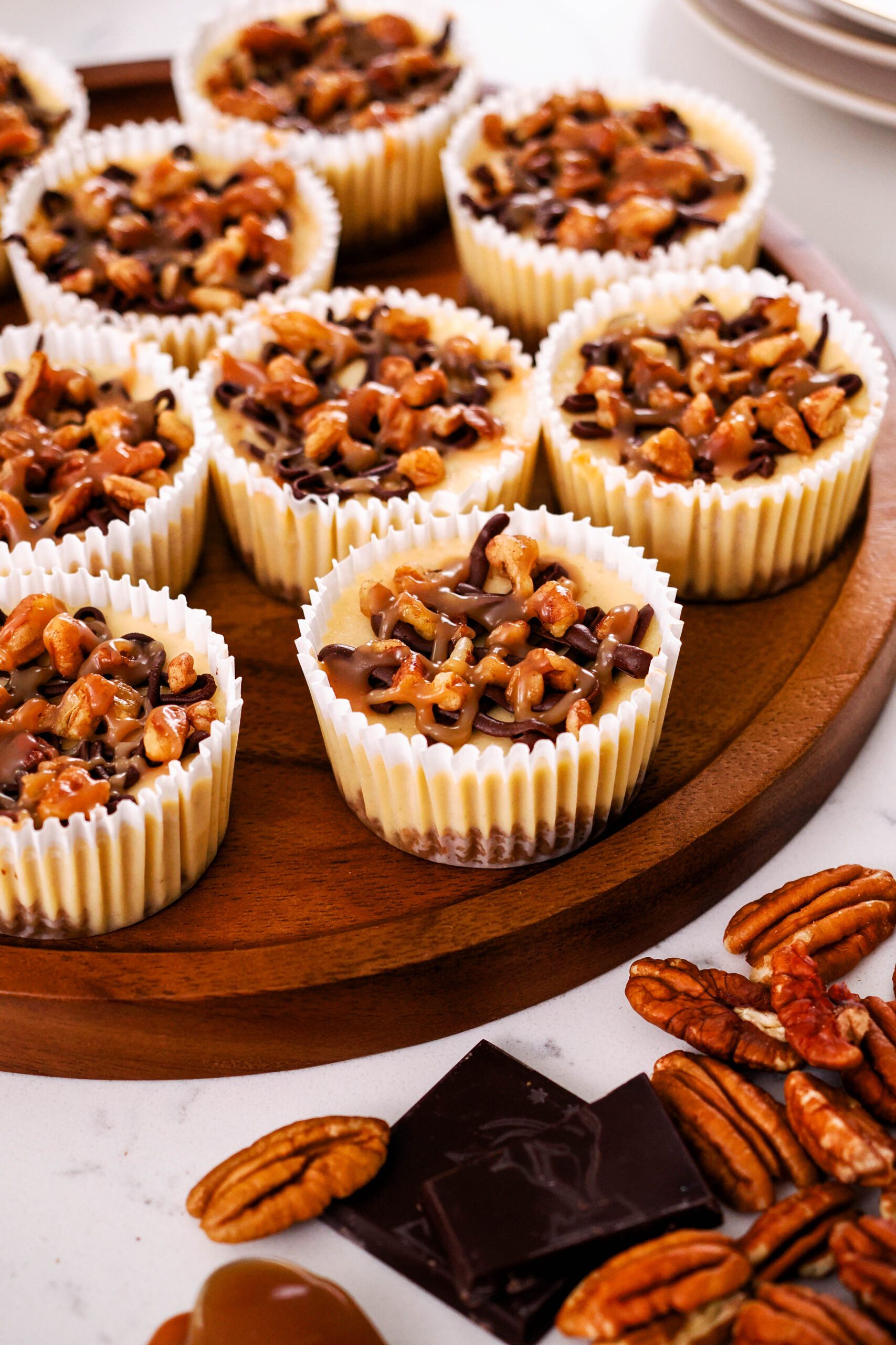 Mini turtle cheesecakes are plated on a wooden platter with pecans and chocolate squares around it.