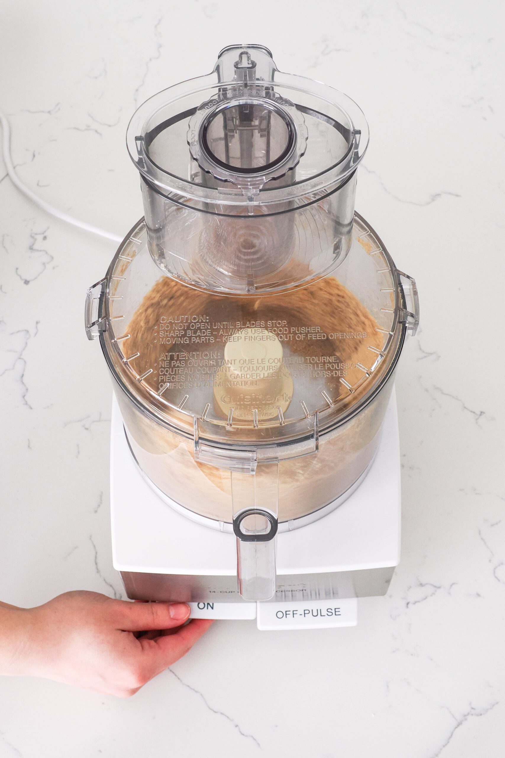 A hand presses the "on" button on a food processor with pecans and graham crackers.