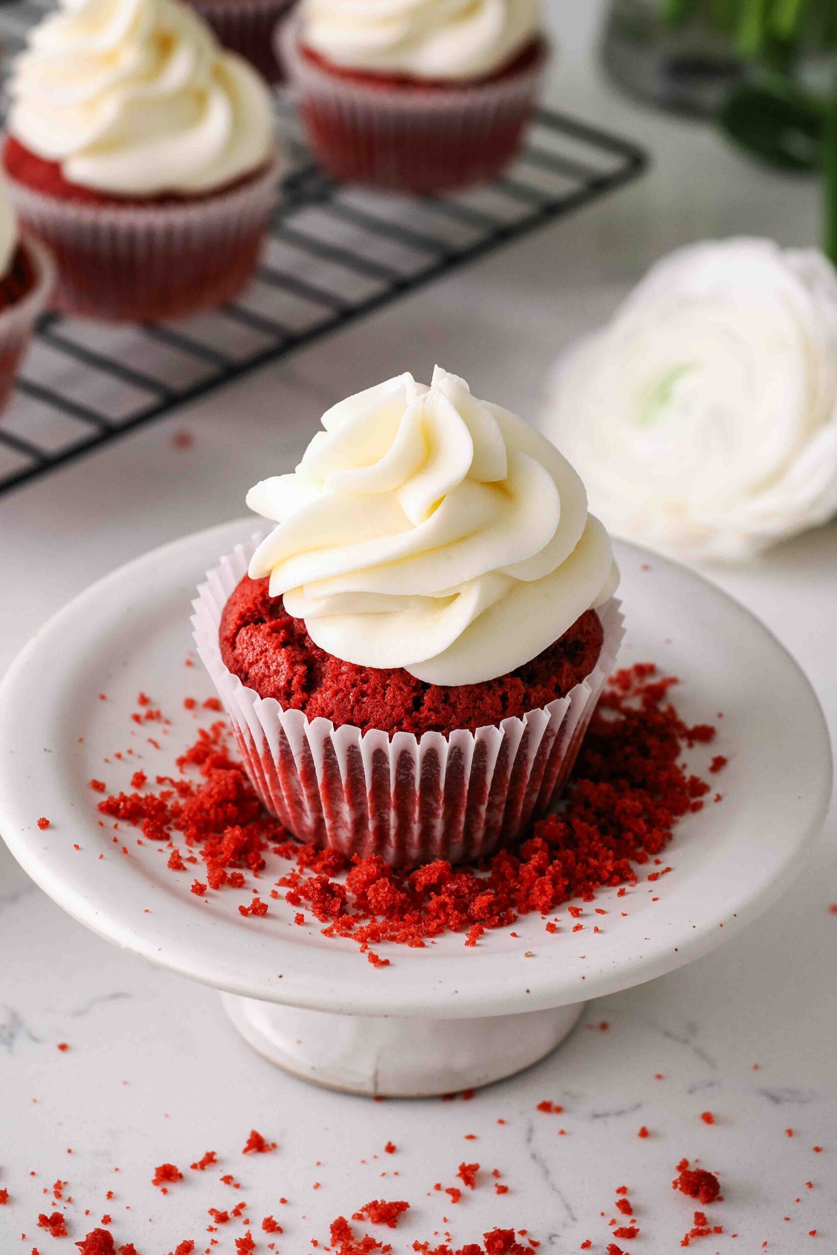 A red velvet cupcake with cream cheese frosting on a cupcake stand with red velvet crumbs.