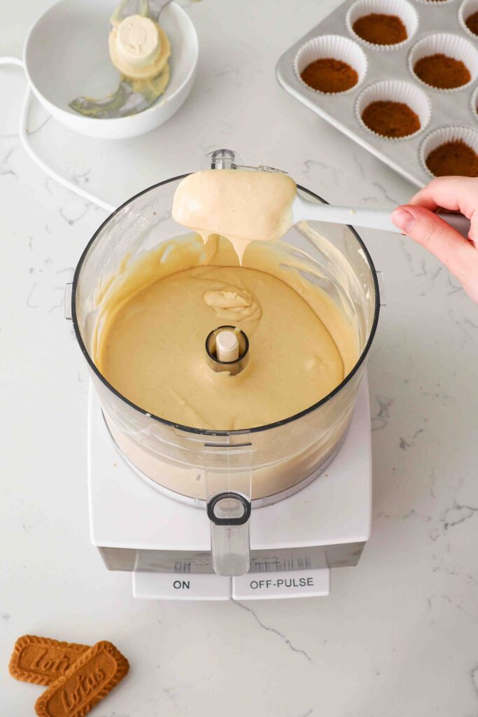 A spatula holds up Biscoff cheesecake batter over a food processor bowl.