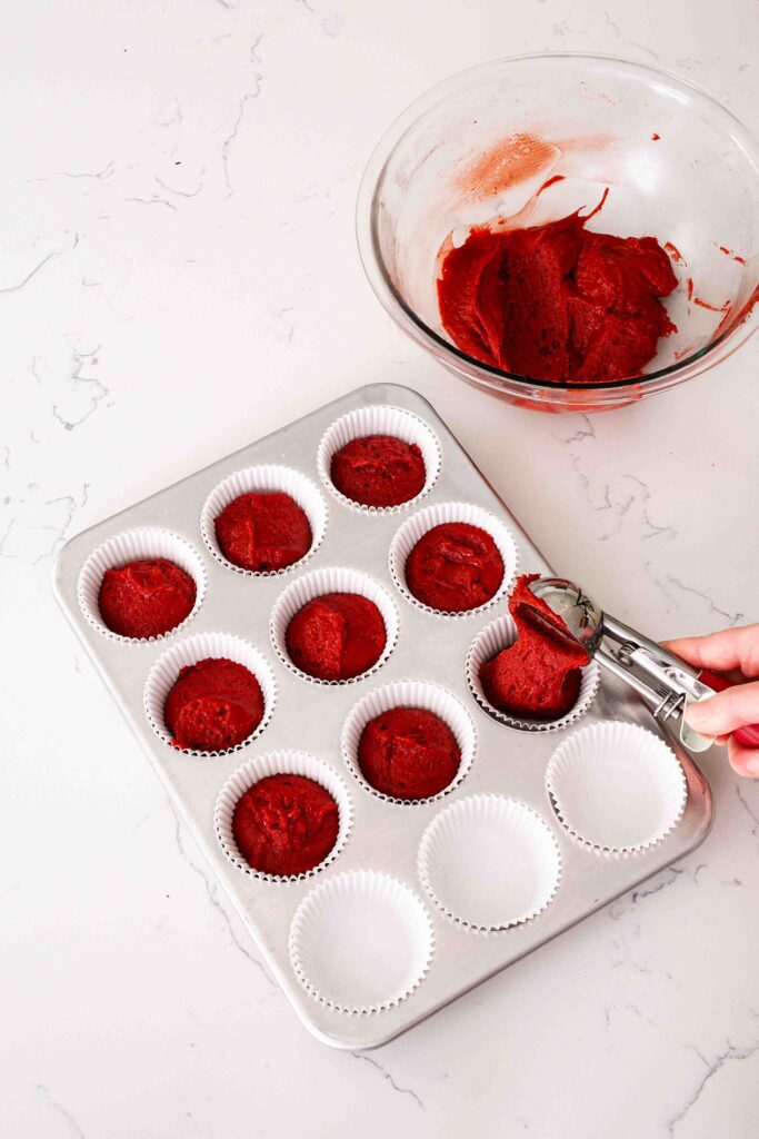 A cookie scoop portions red velvet cake batter into paper cupcake liners.