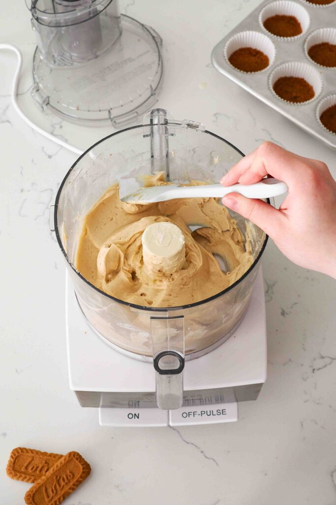 A spatula scrapes down the side of a food processor bowl with Biscoff cheesecake filling on the sides.