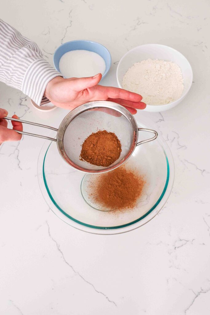 Two hands sift cocoa powder into a glass bowl.
