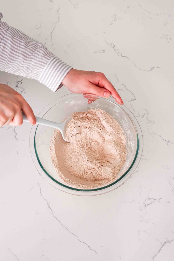 Two hands use a spatula to stir cocoa powder into dry ingredients in a glass bowl.