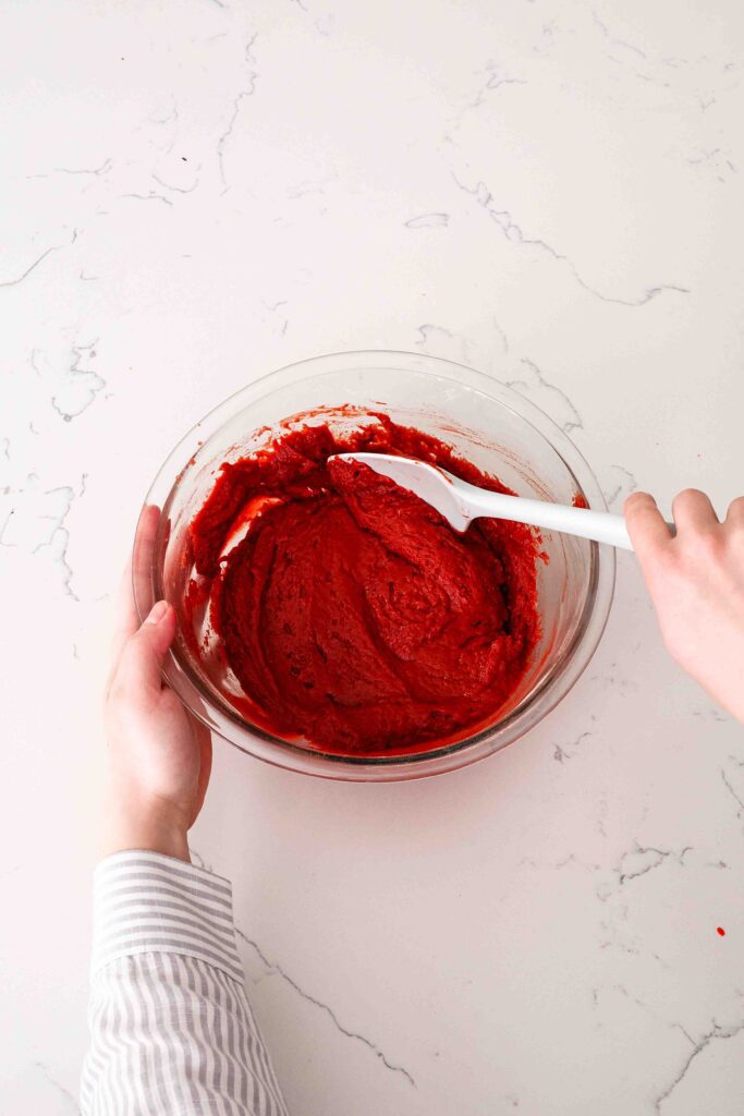 Two hands fold together the red velvet cake batter in a glass bowl.