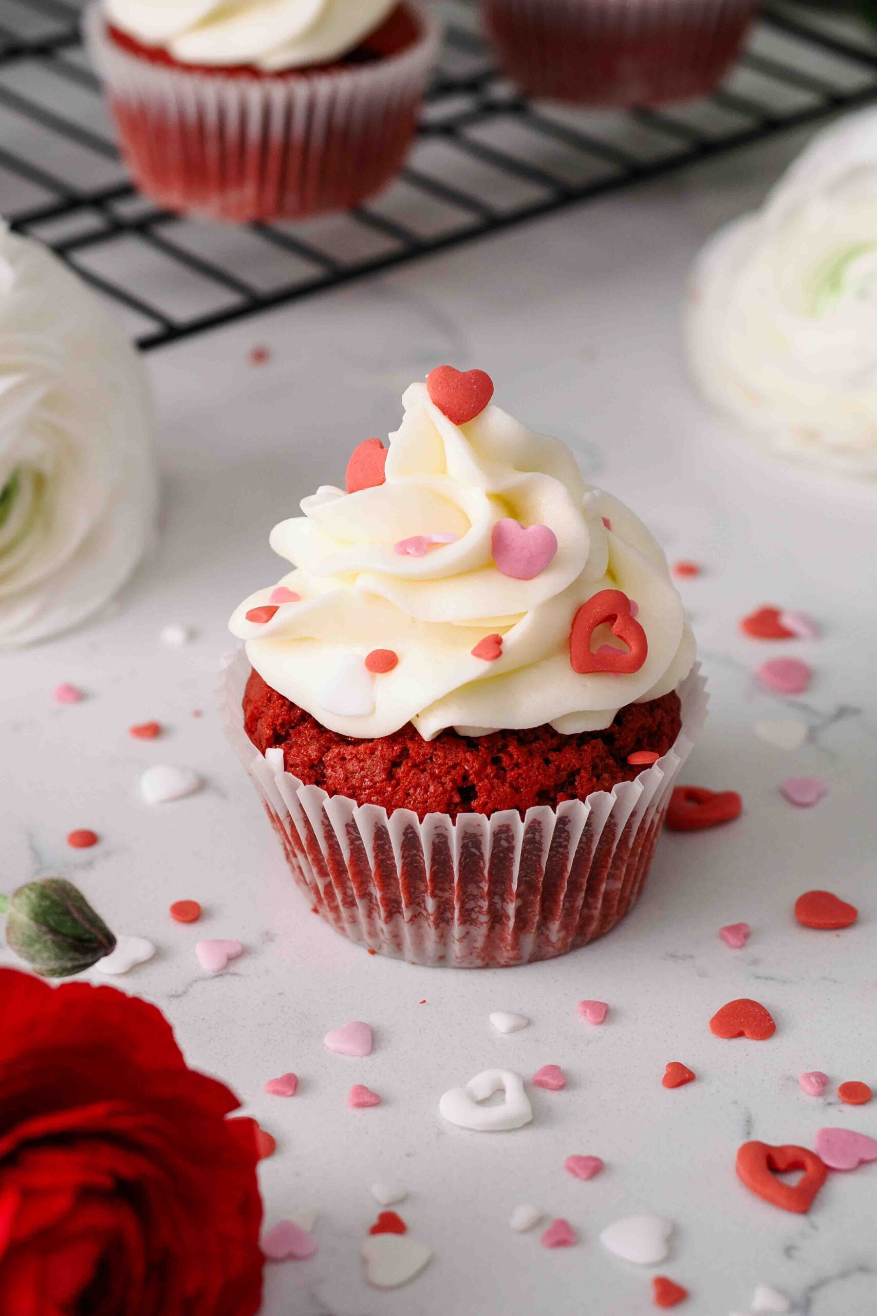 A red velvet cupcake is topped with red, pink, and white heart sprinkles near a red ranunculus flower.