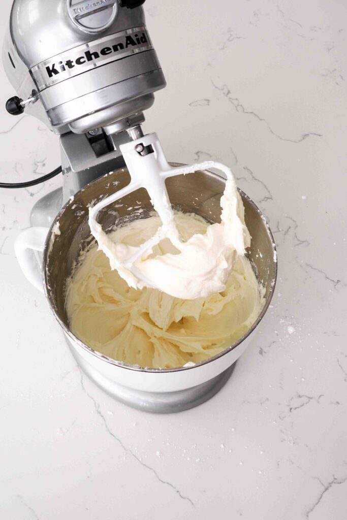 A thick cream cheese frosting is clumped in the base of a beater attachment on a stand mixer.