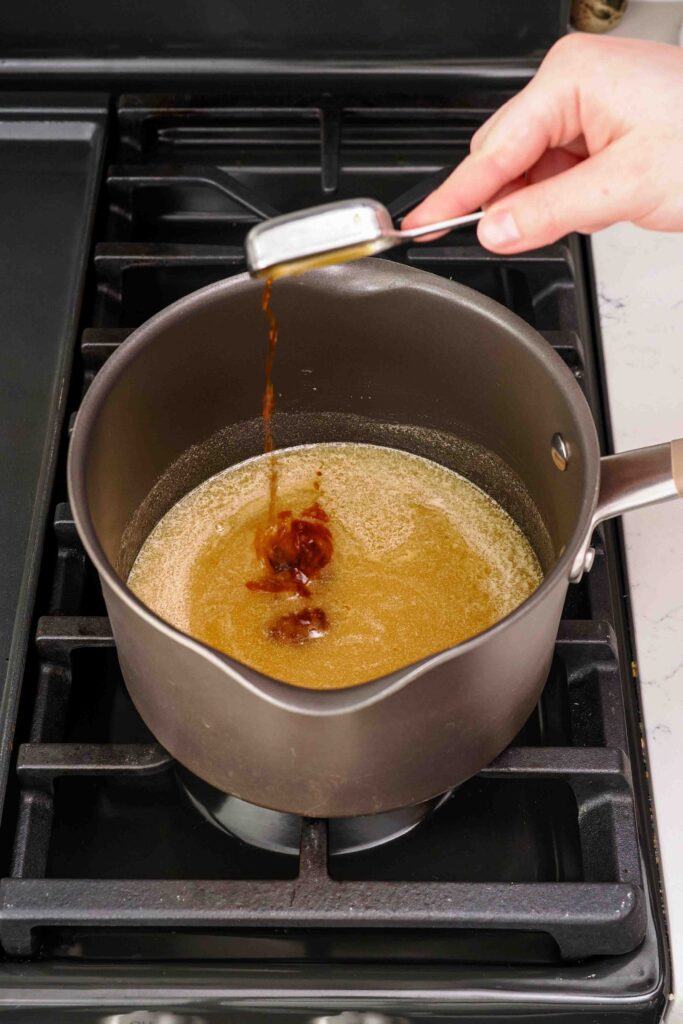 A hand pours vanilla extract into a pot with honey syrup.