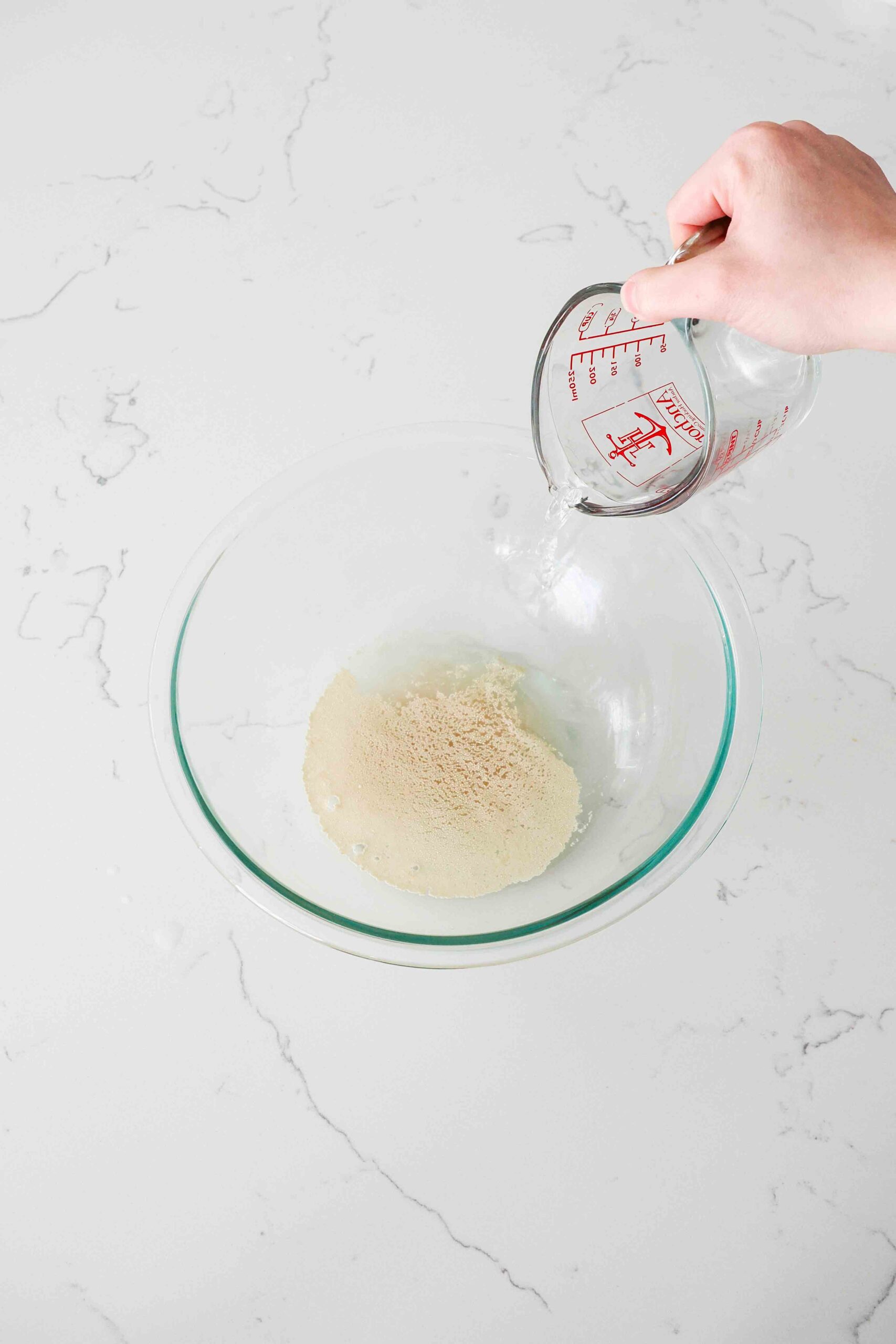 A hand pours warm water from a measuring cup over yeast in a glass mixing bowl.