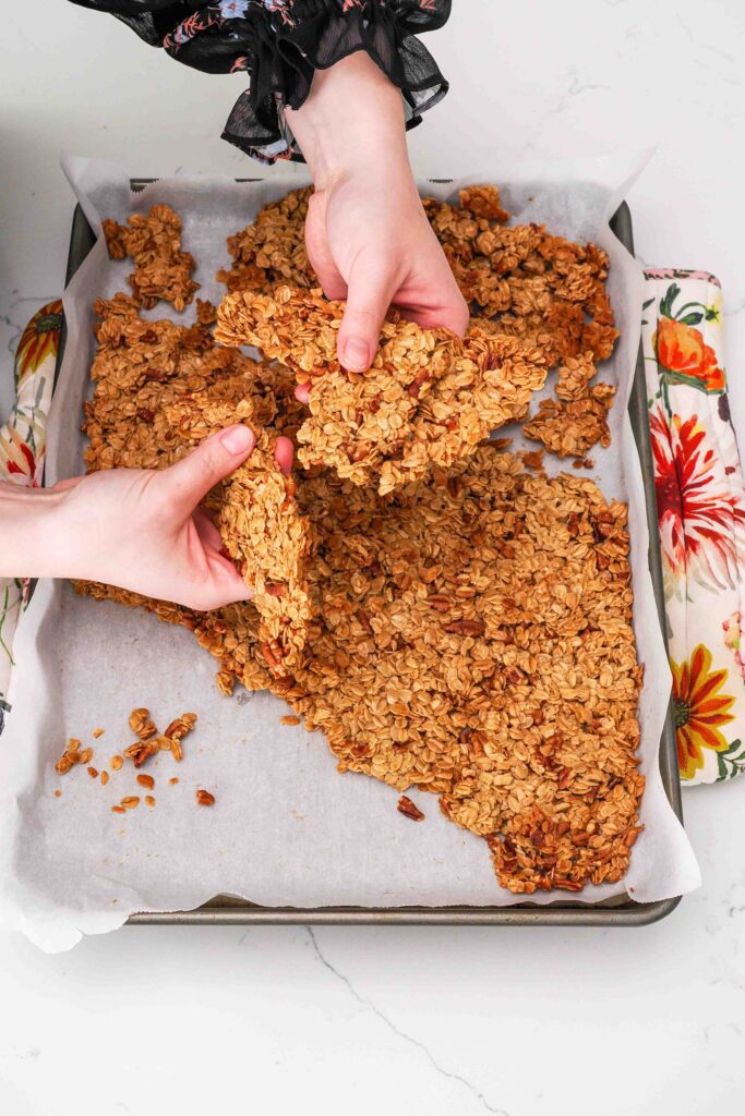 Two hands break up a large clump of granola over a parchment-lined baking sheet.