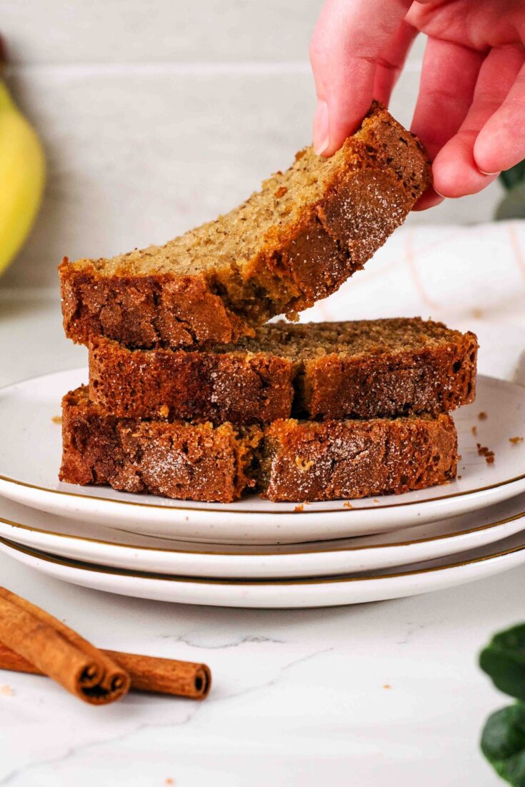 A hand takes a slice of cinnamon banana bread from a stack on a plate.