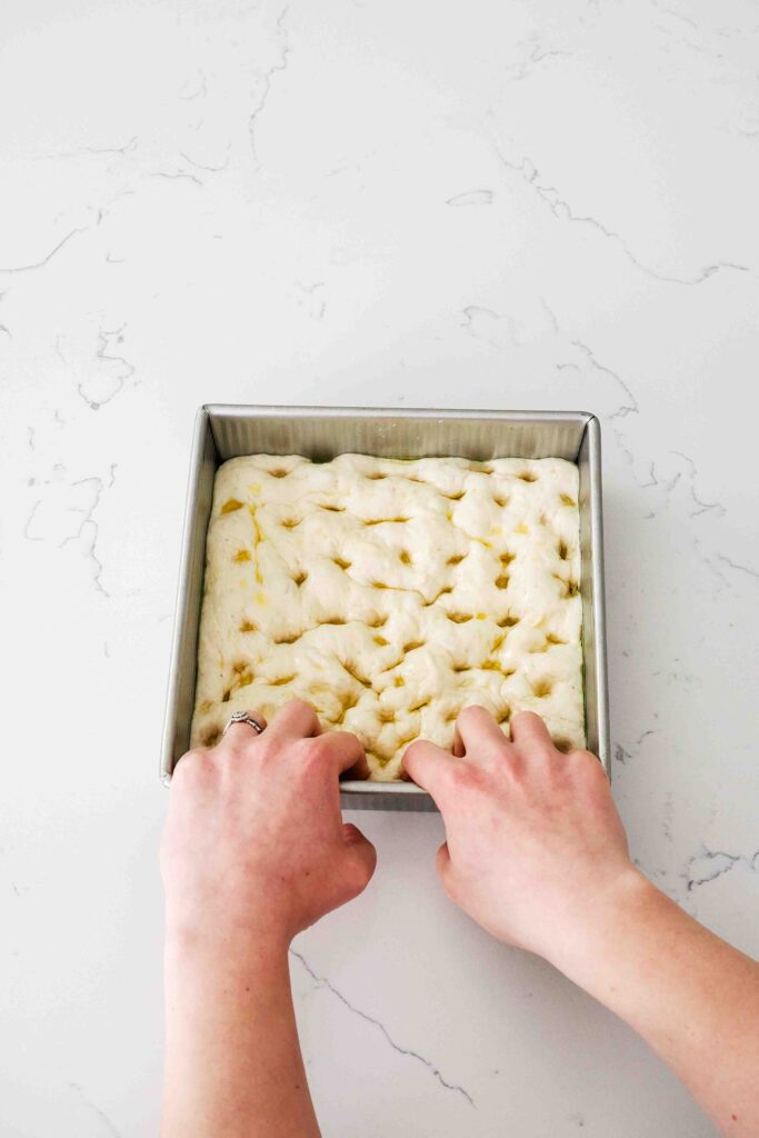 Two hands press their fingertips into proofed focaccia dough in a square pan.