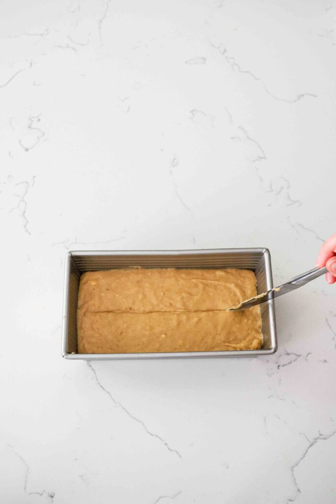 A hand draws a line with a butter knife through batter in a loaf pan.